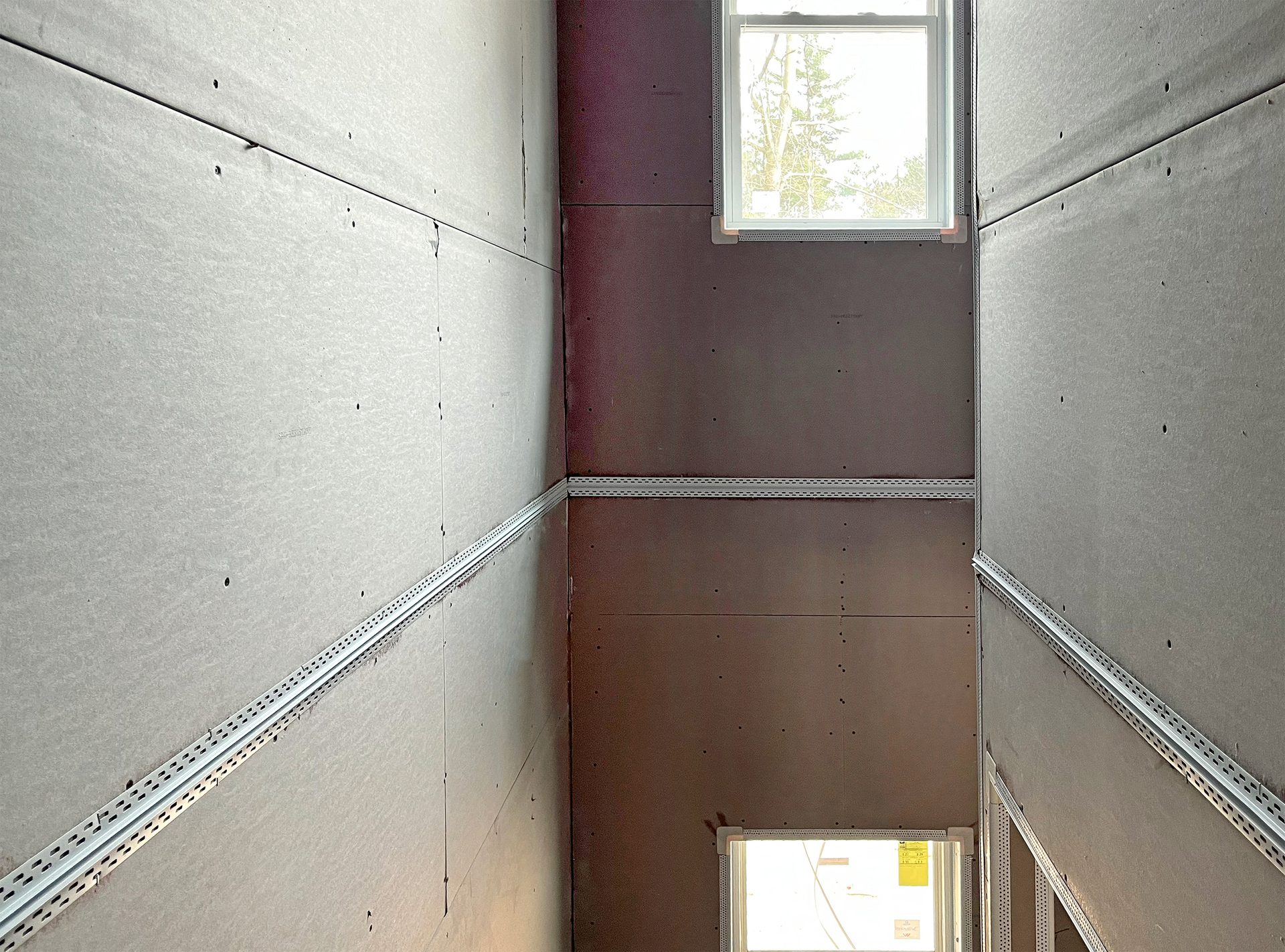 Room under construction with gray and brown drywall panels, metal joint strips, and windows.