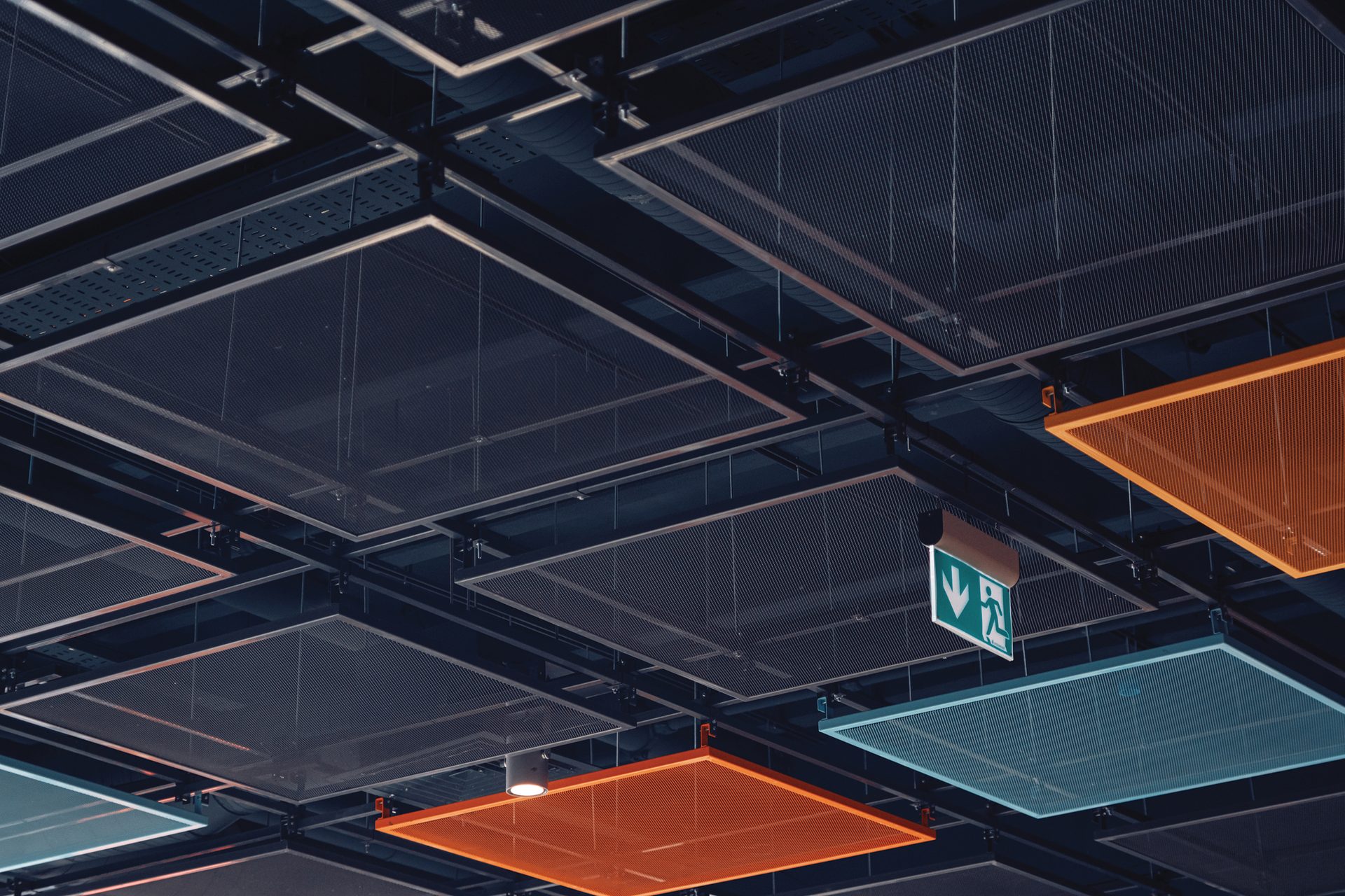 Modern ceiling with square mesh panels, some orange and blue, and an emergency exit sign.