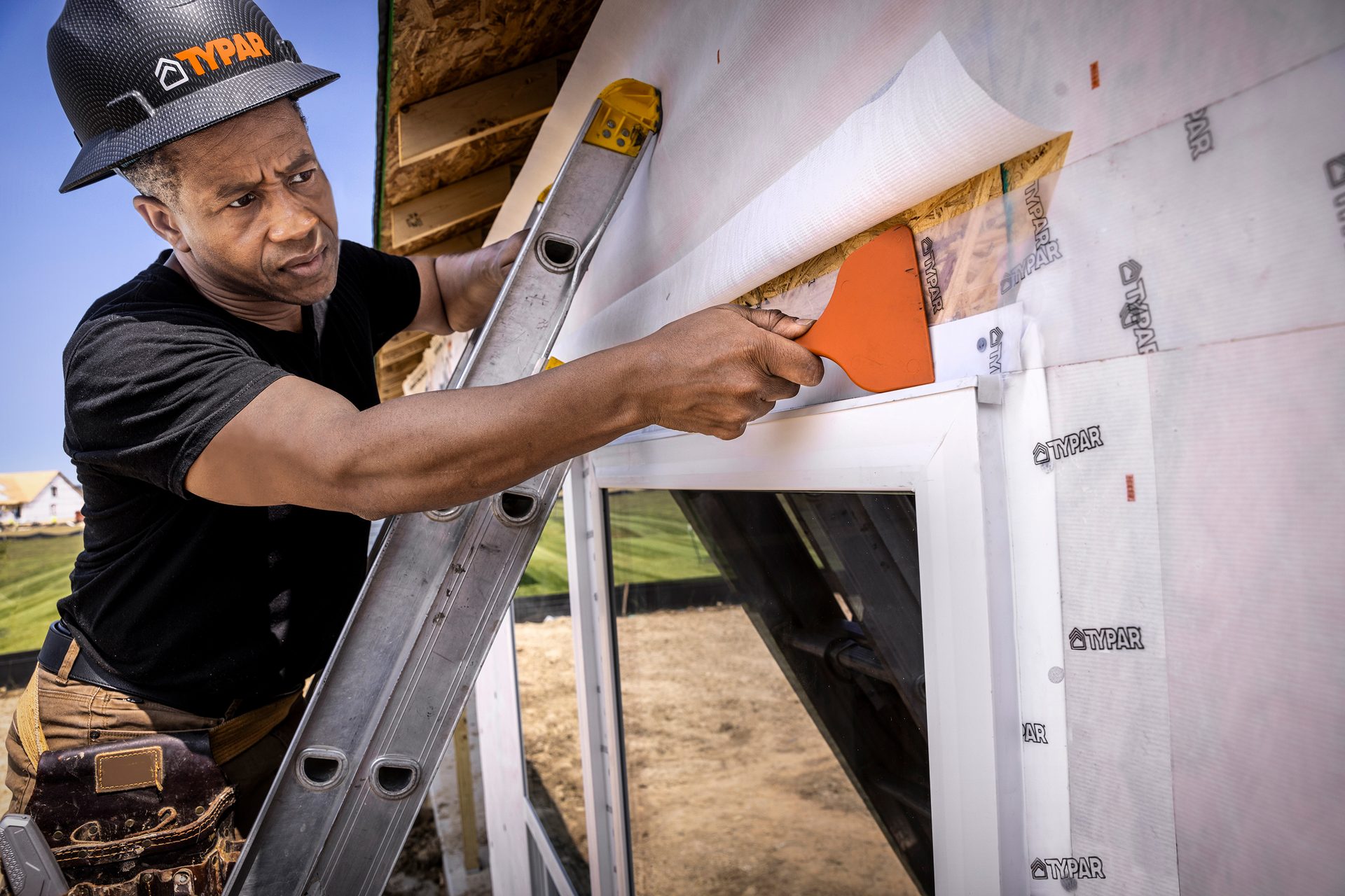 Man on ladder applying TYPAR house wrap around a window with an orange tool.
