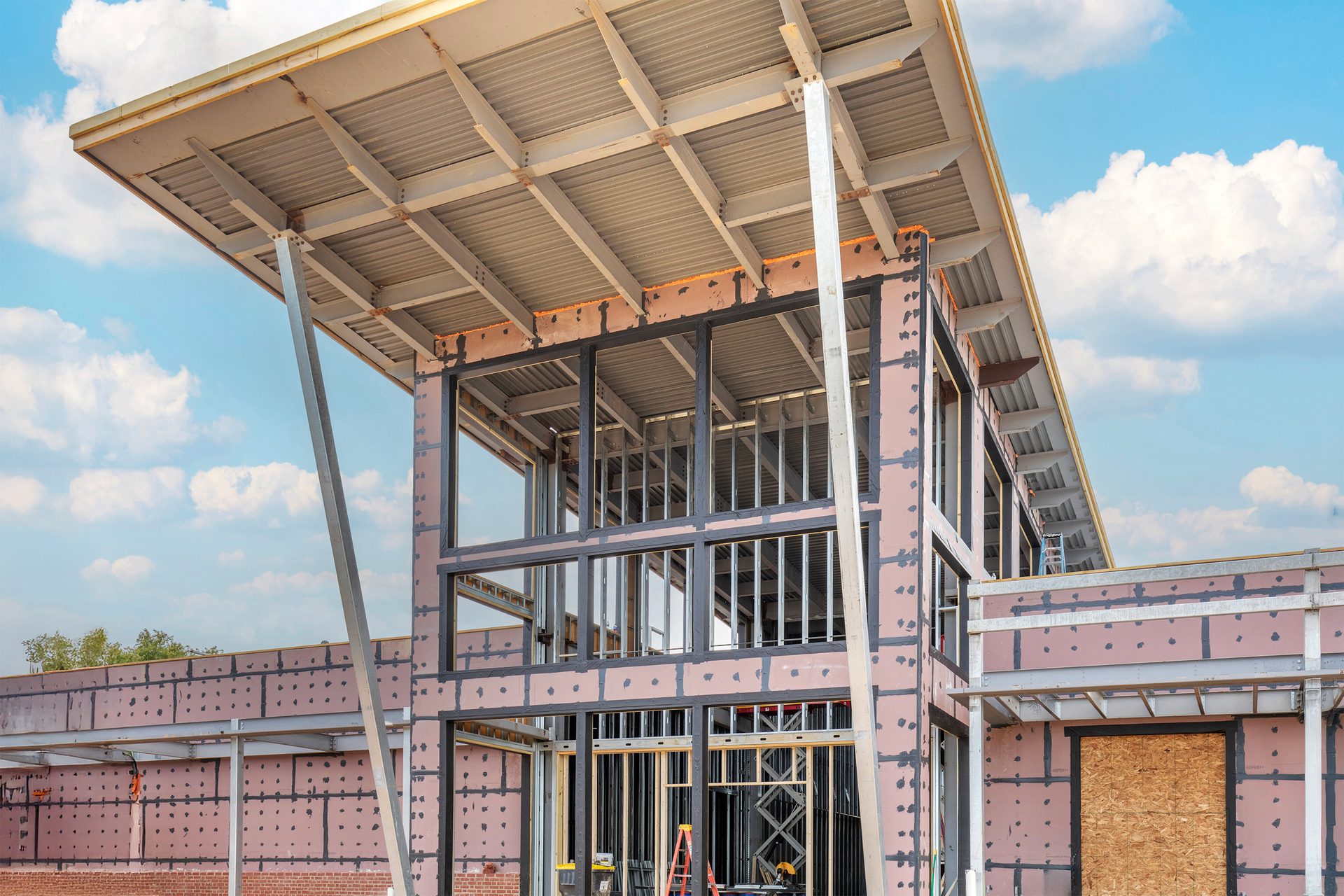 Construction site of a modern building with an exposed steel frame, large angled roof, and pink insulation.