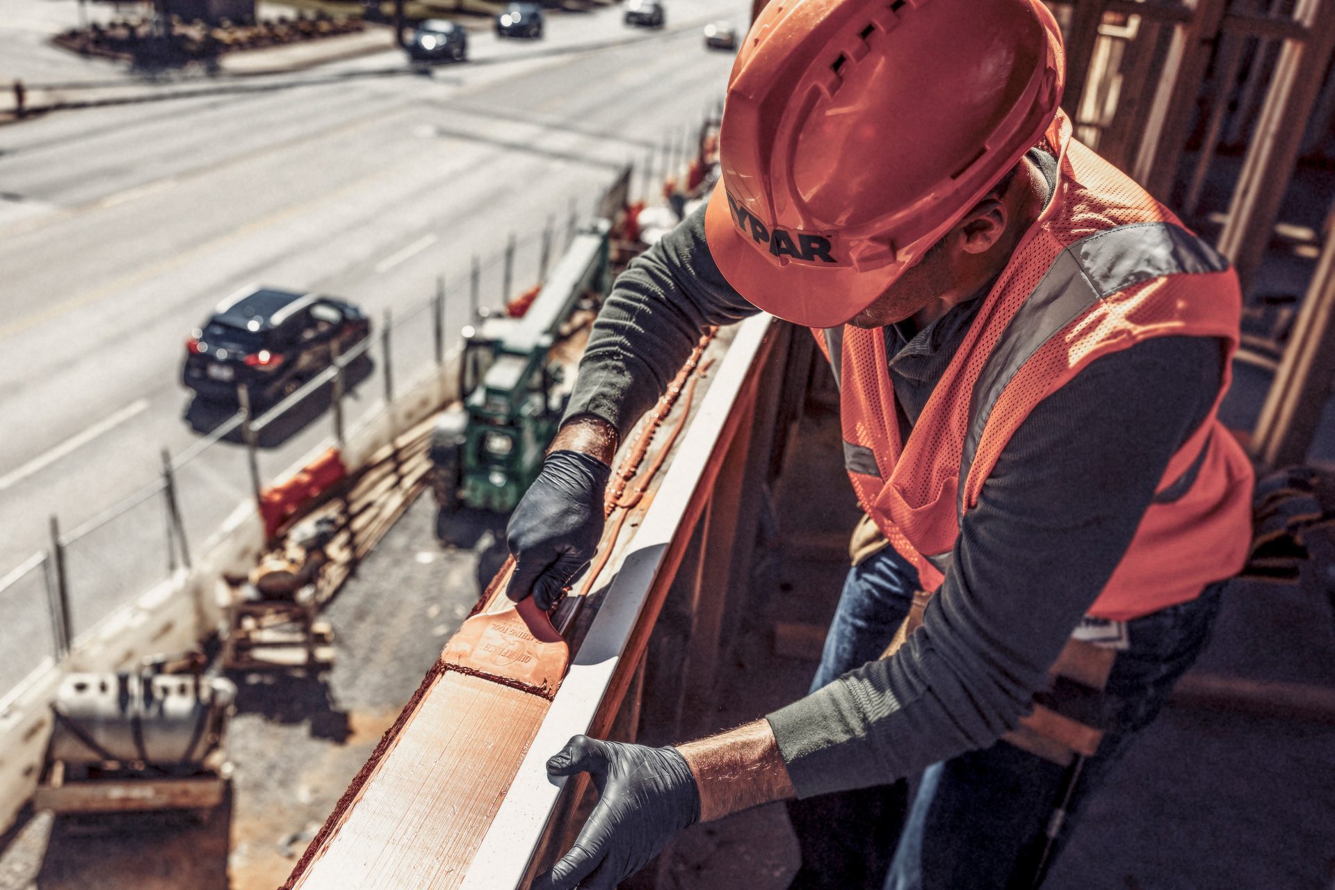 Construction worker in hard hat and safety vest applies sealant to a beam overlooking a road.