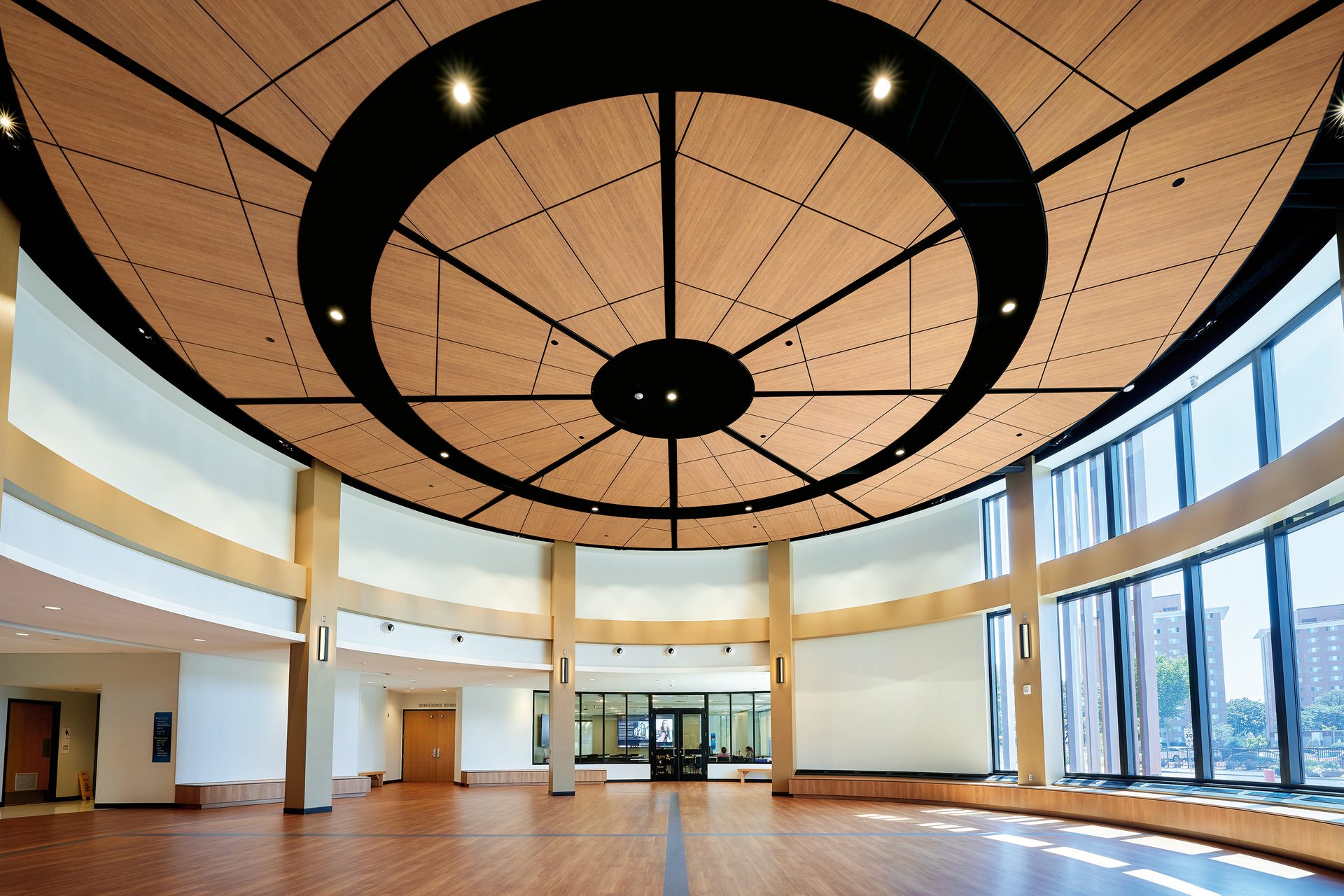 Modern circular hall with a radial wooden ceiling, warm lights, and tall windows.