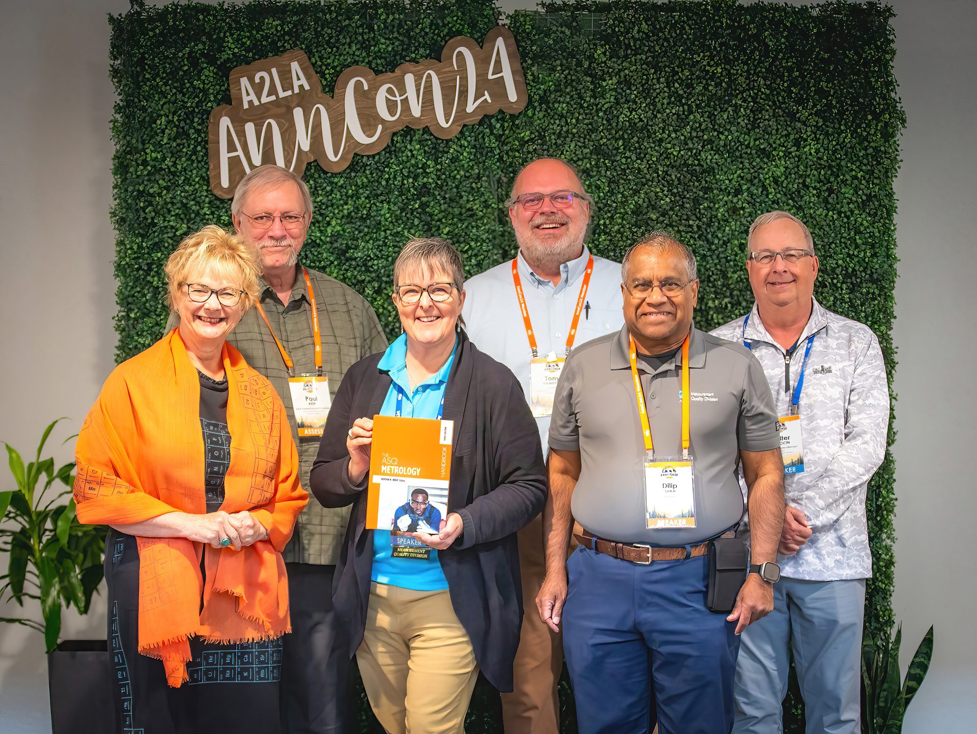 Six people, three women and three men, posing in front of a green wall with "A2LA AnnCon 24" written on it. One woman is holding a book.