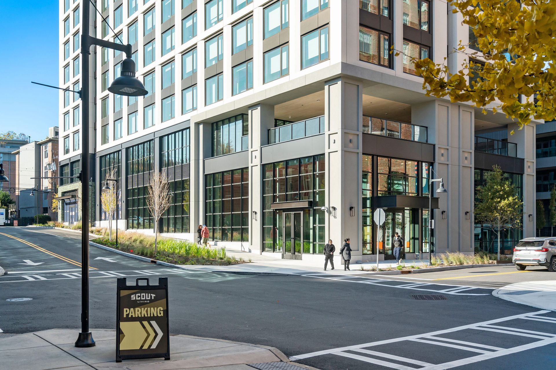 Modern building at a street corner, with people, a car, and a "Scout Living Parking" sign.