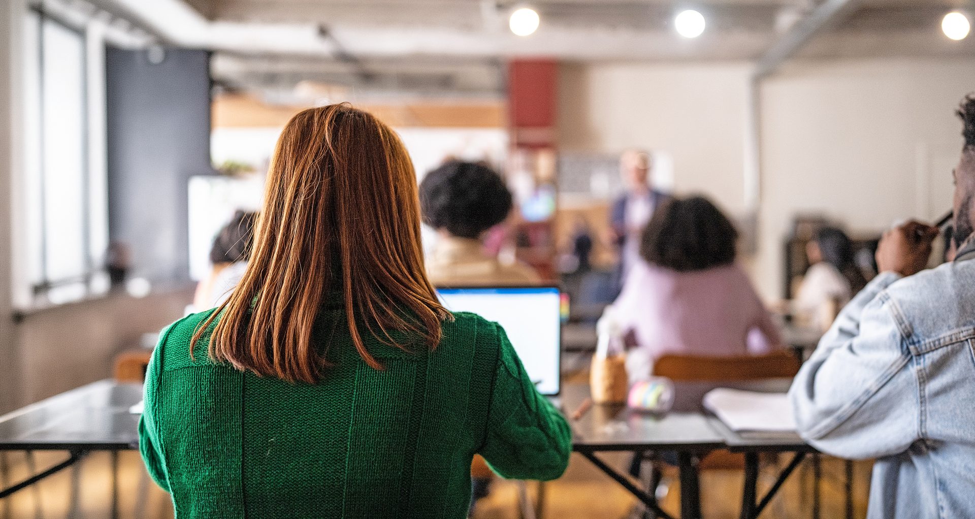 A person with a laptop, seen from behind, attending a class with other students.