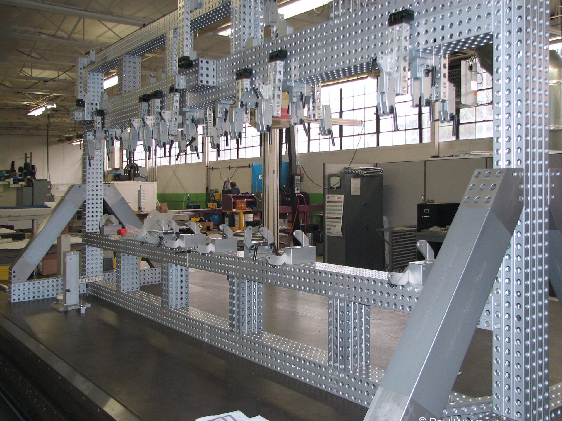 Large, modular perforated metal jig with numerous clamps in a workshop setting.