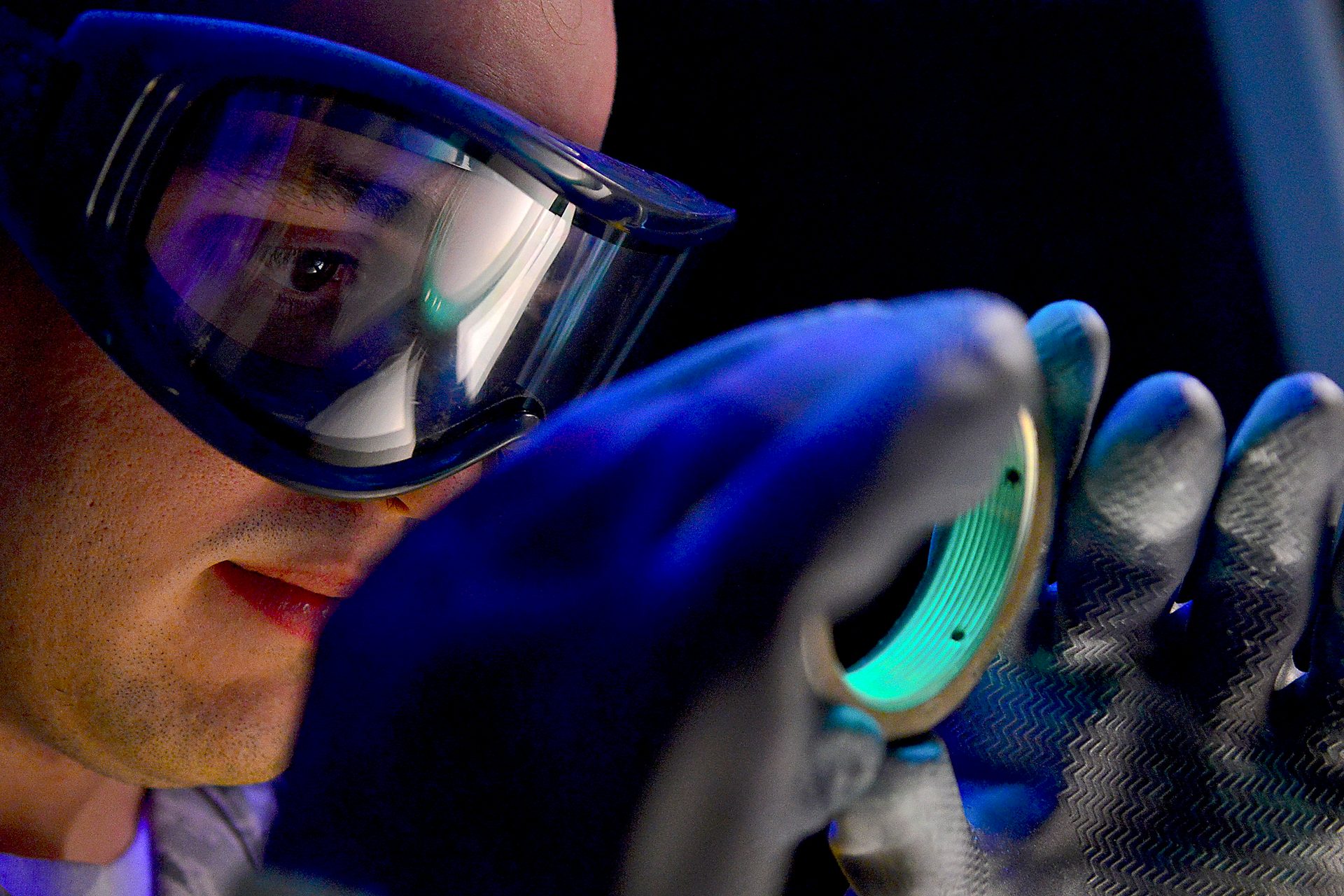 Person in safety goggles examining a glowing green ring held in a gloved hand.
