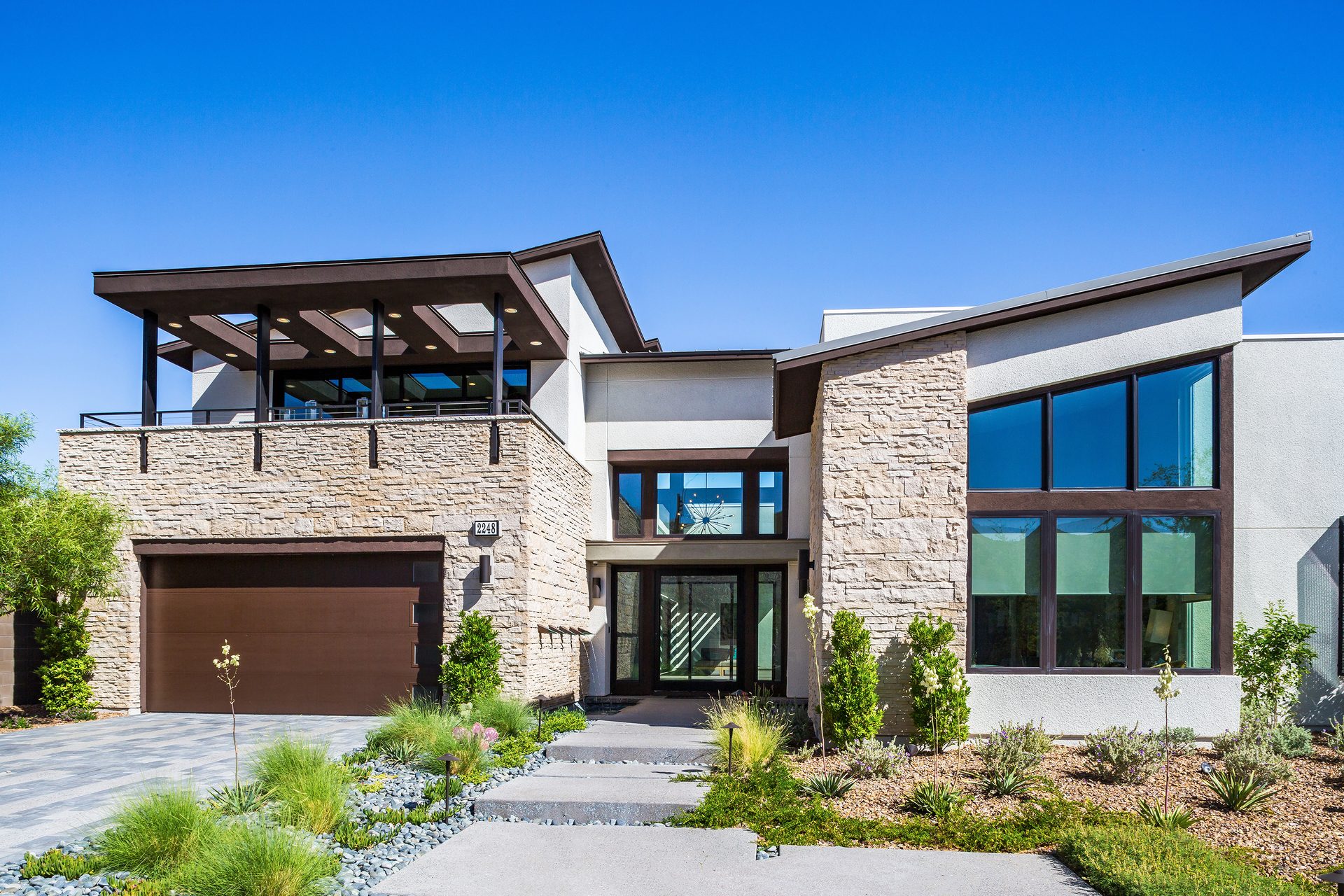 Modern luxury house exterior with stone and stucco, large windows, balcony, garage, and desert landscaping.