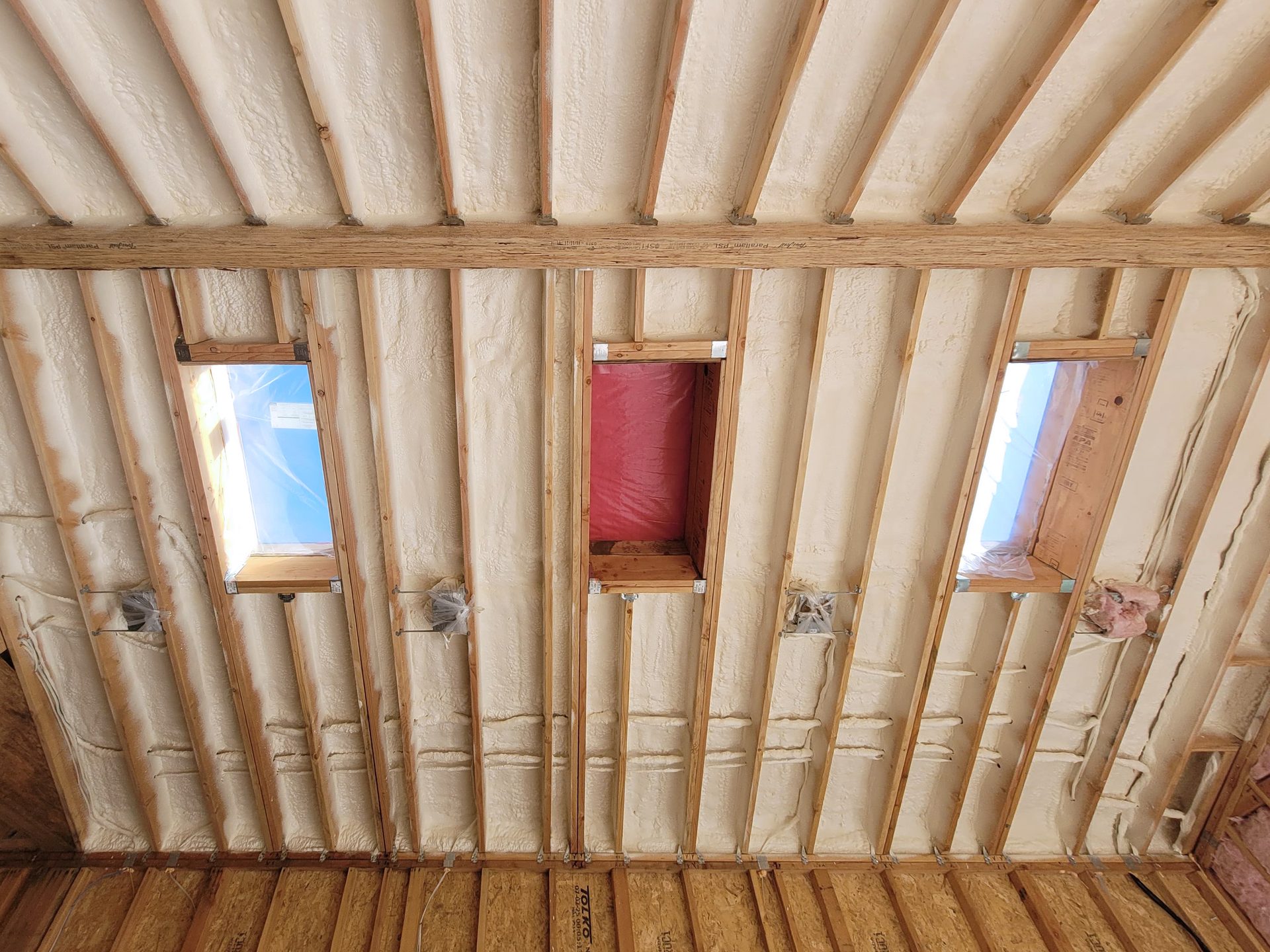 Spray foam insulation covers a vaulted ceiling with exposed wooden rafters and three skylight openings.
