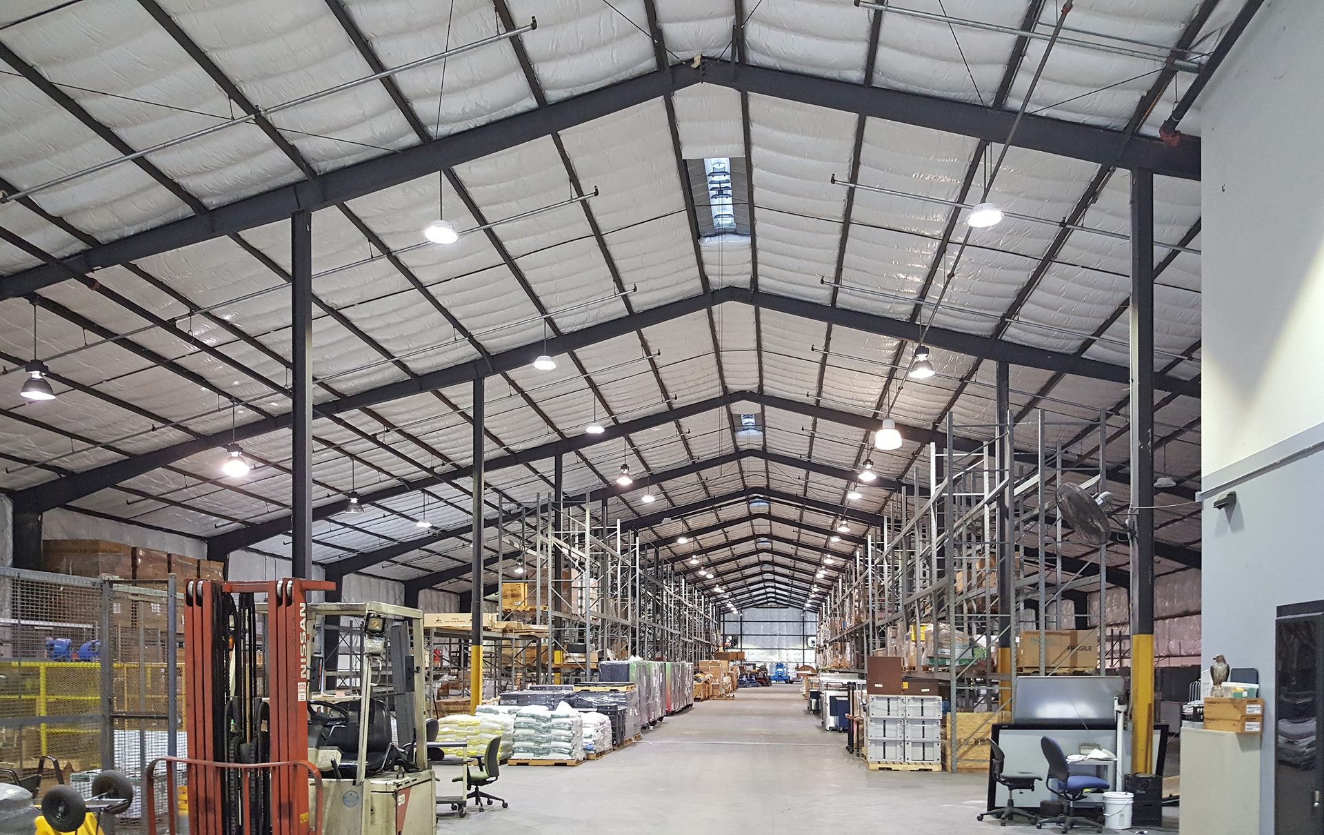 Large warehouse with high ceilings, structural beams, rows of shelving, forklifts, and office areas.