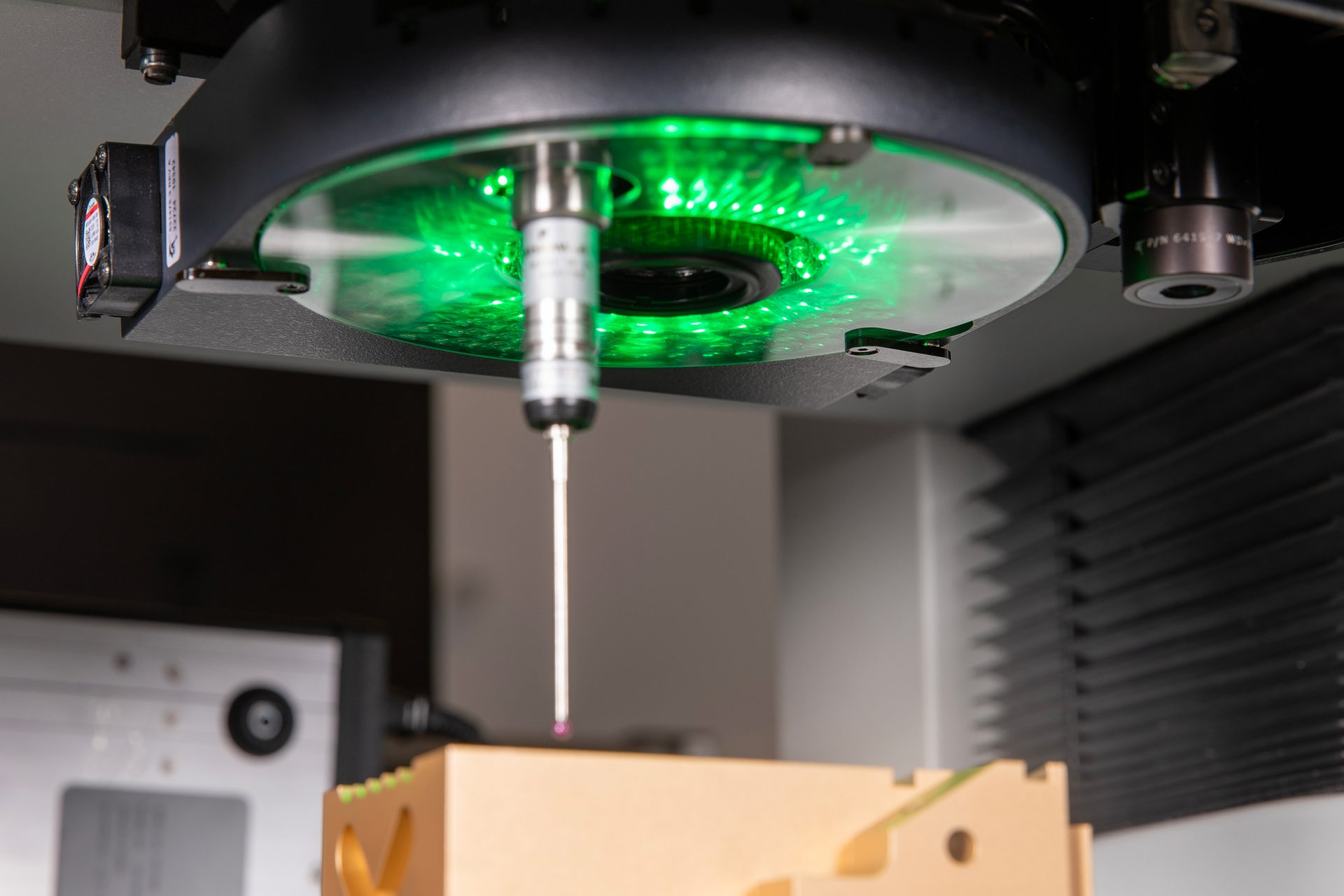 A precision measuring machine (CMM) probe measures a part, illuminated by green light.