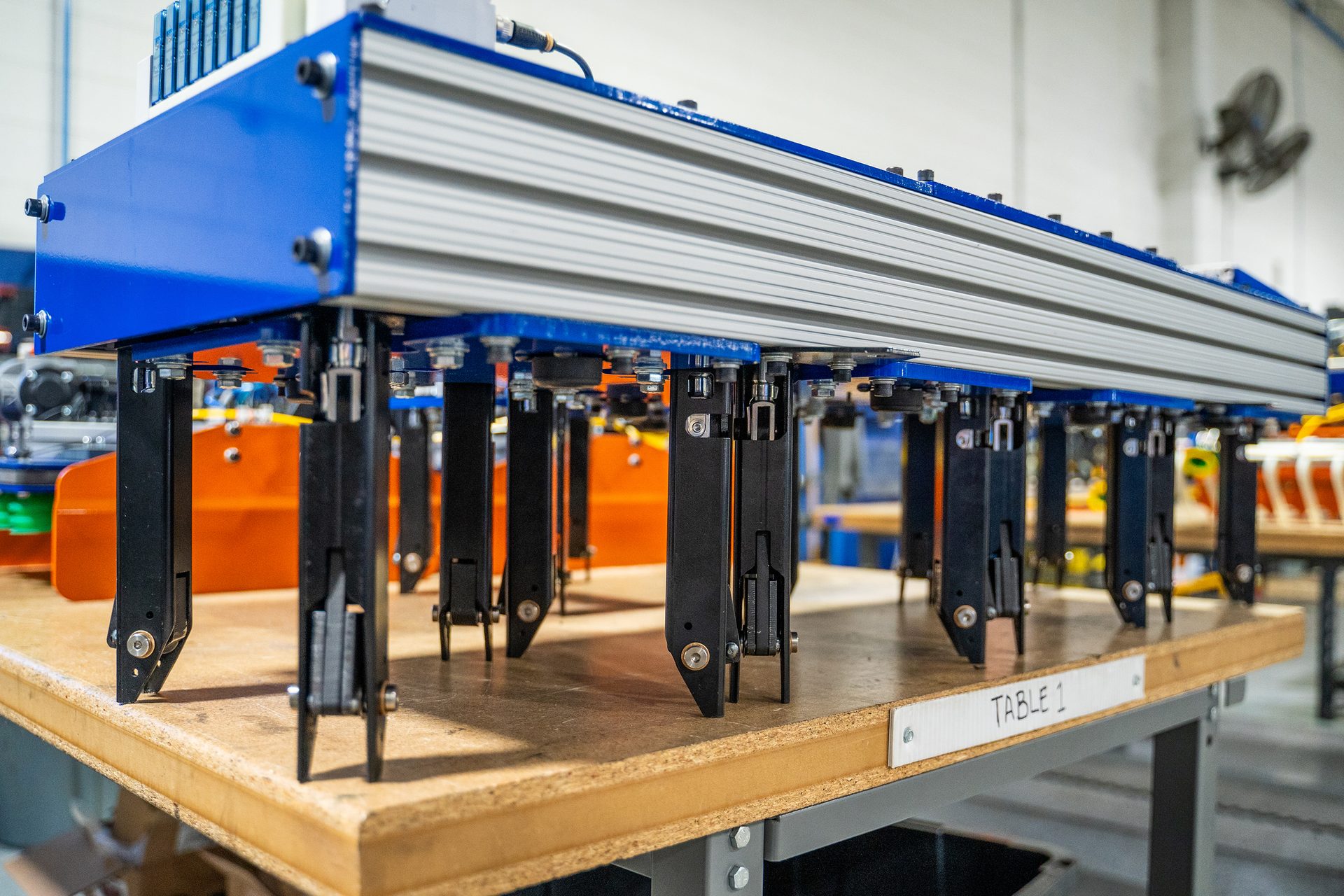 A blue and silver machine with multiple black grippers rests on a wooden table labeled "TABLE 1".