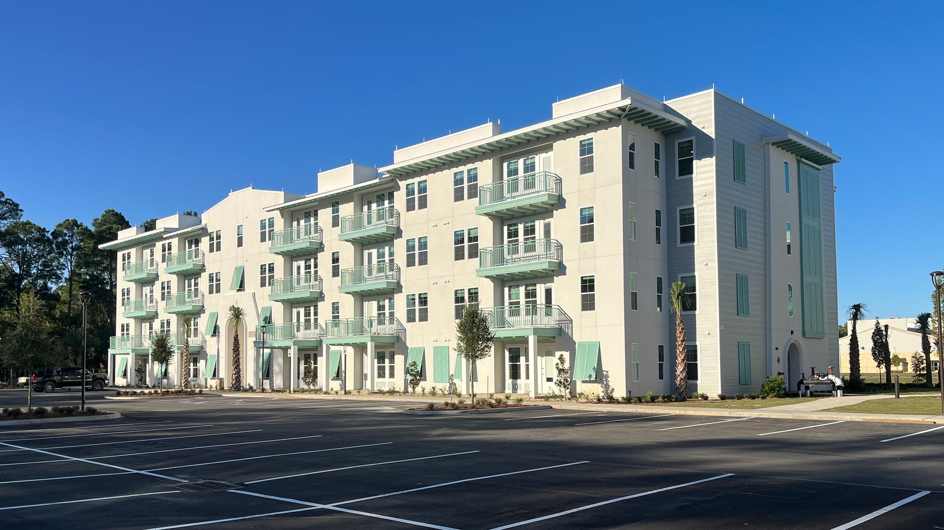 White apartment building with mint green balconies and shutters, an empty parking lot, and a clear blue sky.