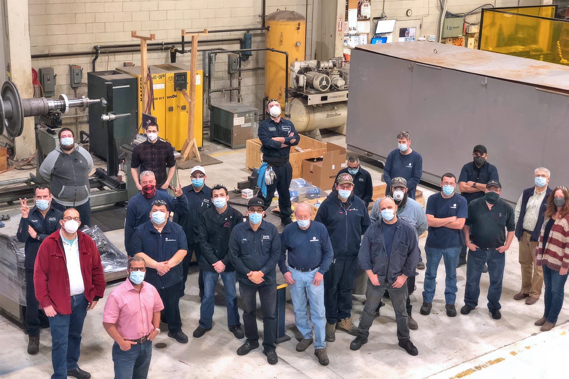Group of masked factory workers and staff pose for a team photo in an industrial workshop.