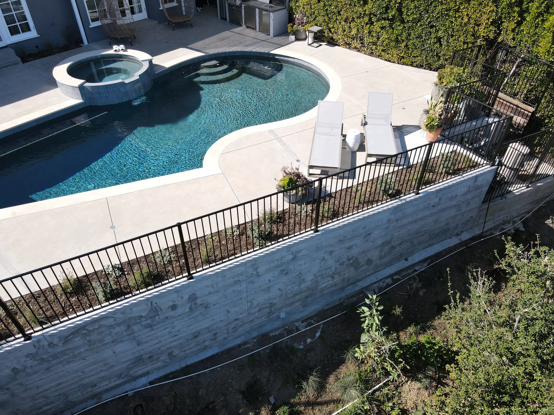 Aerial view of a modern backyard with a dark pool, hot tub, concrete patio, and a retaining wall.