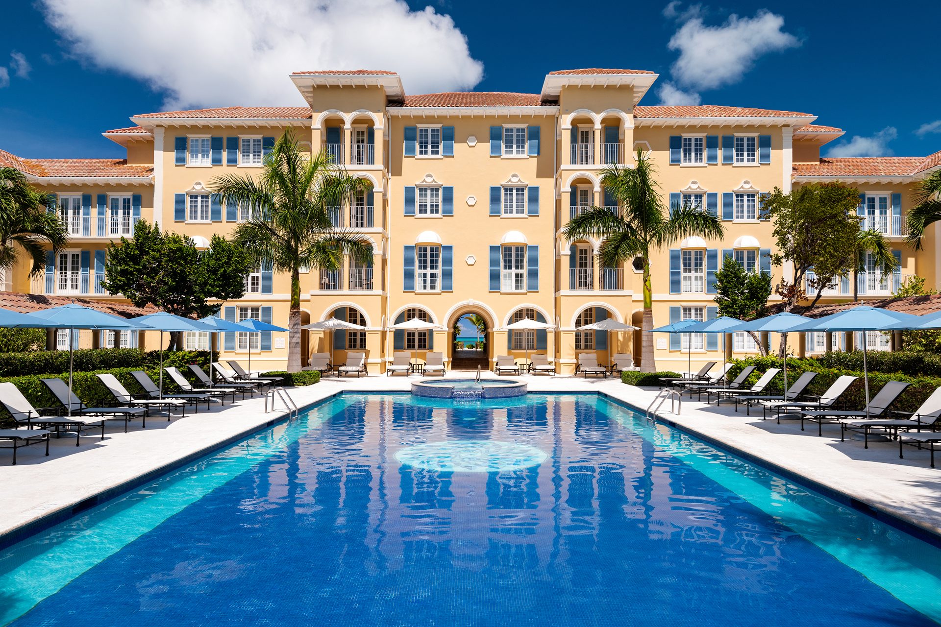 A grand yellow hotel with blue shutters, a mosaic pool, lounge chairs, and an archway revealing the ocean.