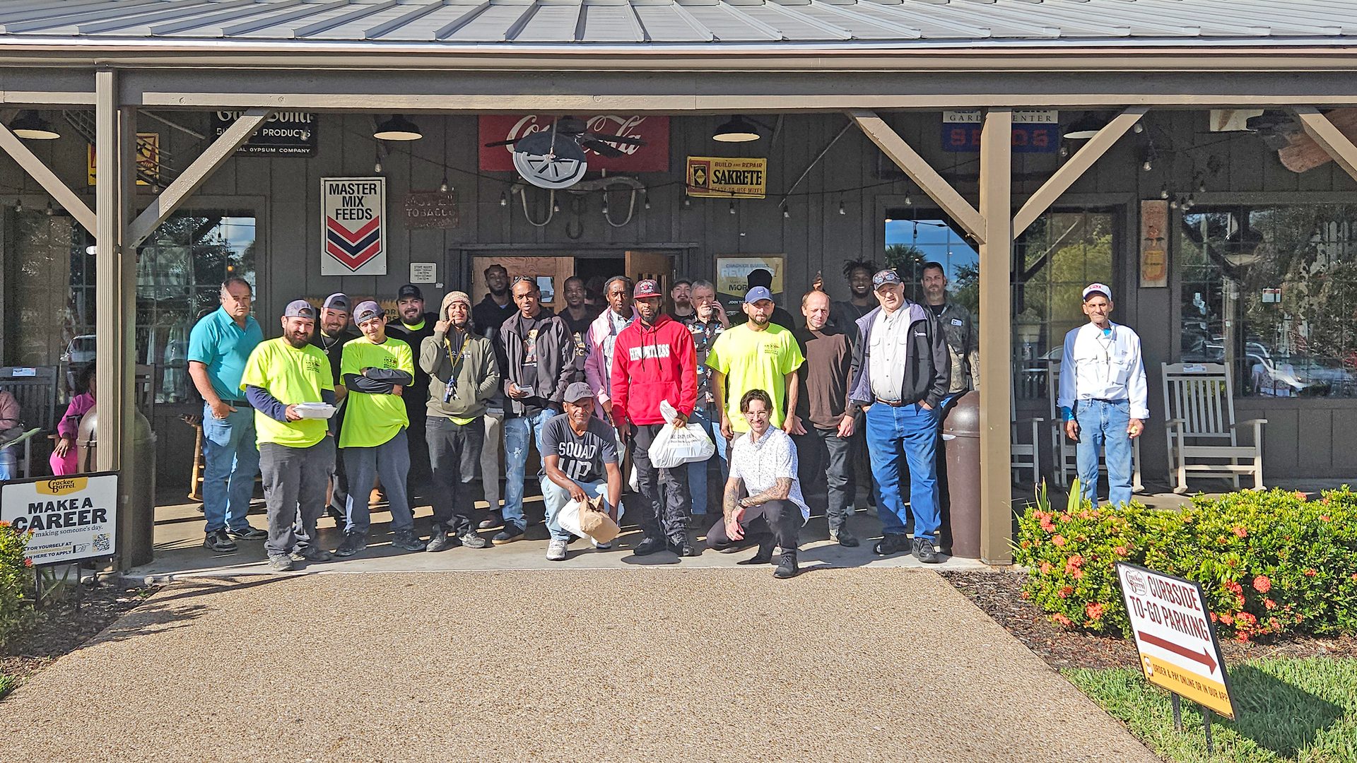 A large group of male workers poses together in front of a restaurant, some holding food containers.