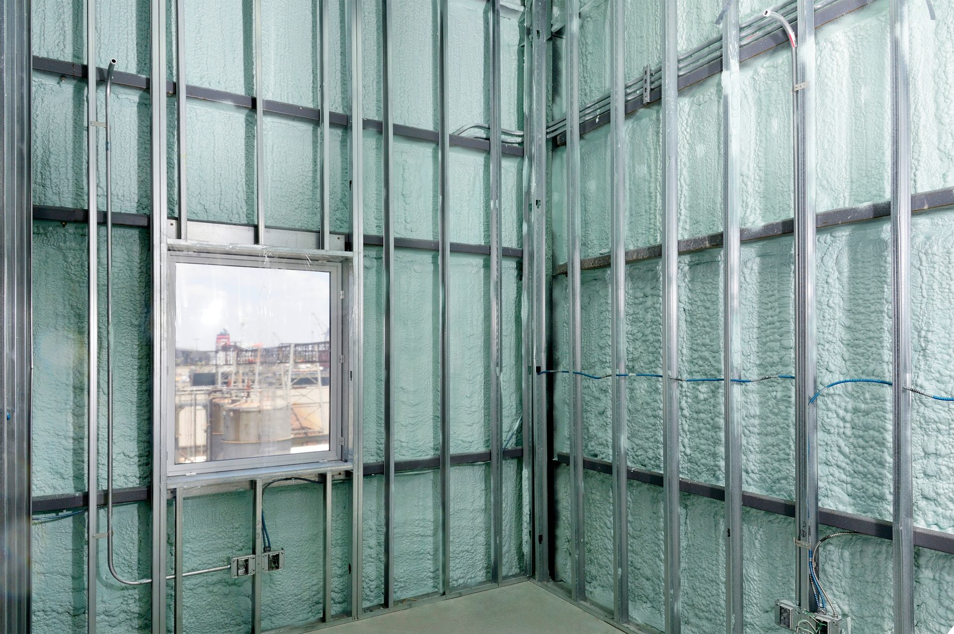 Building interior with metal studs, green spray foam insulation, and a window showing an industrial view.