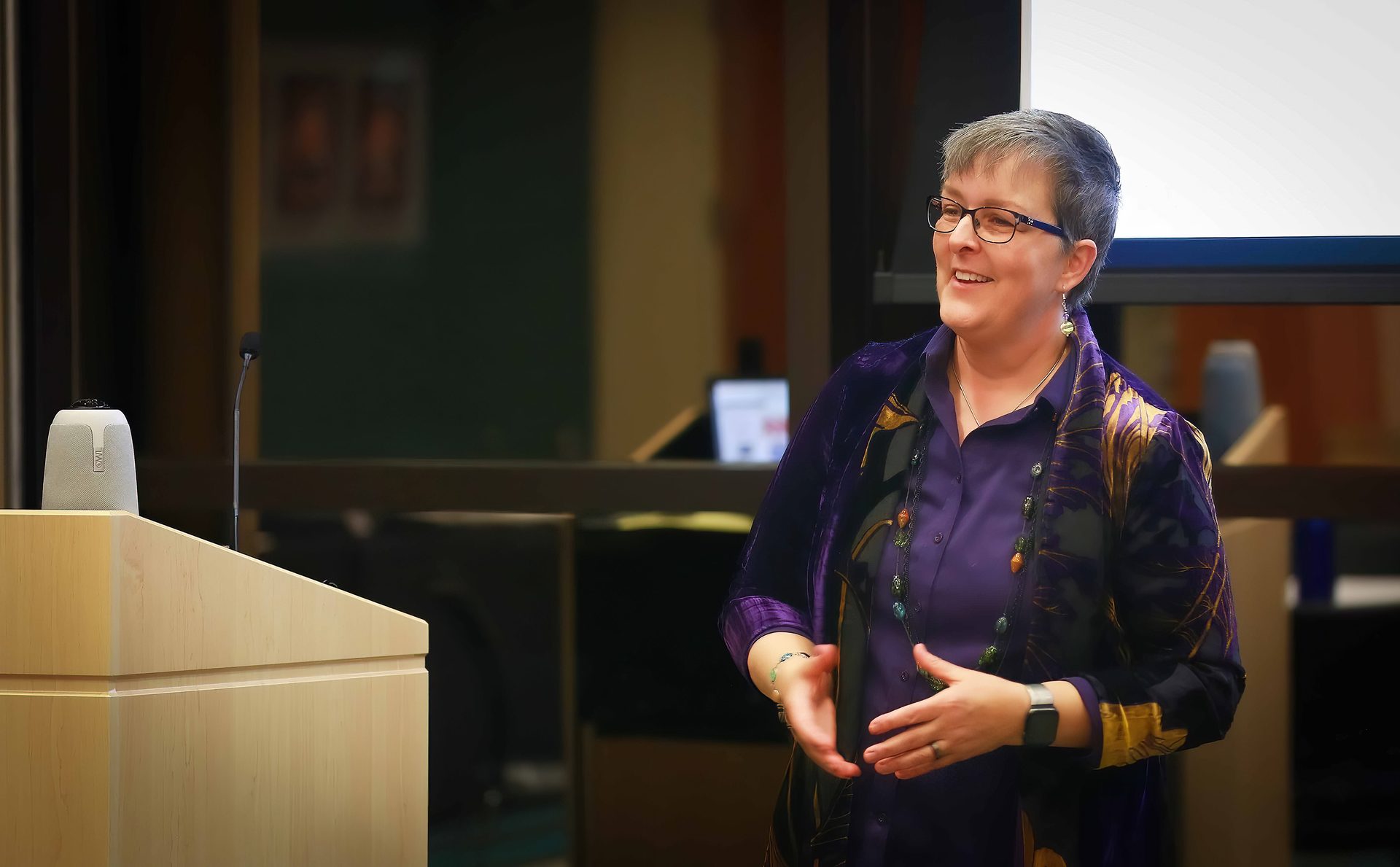 A smiling woman with short gray hair and glasses, wearing a purple shirt and jacket, speaks at a podium.