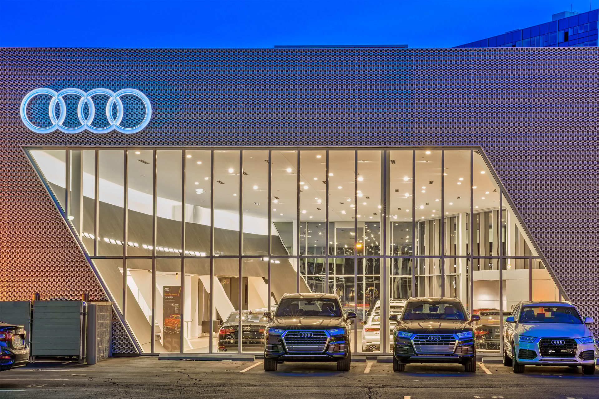 Modern Audi dealership at dusk with a glowing logo, glass facade, and new cars parked outside.