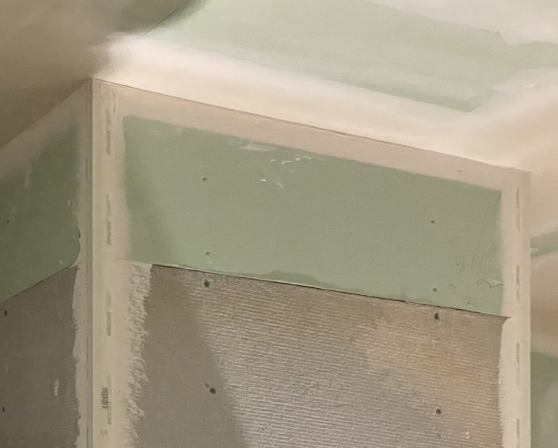 Drywall installation in progress, showing green and gray boards, taped seams, and screw holes.