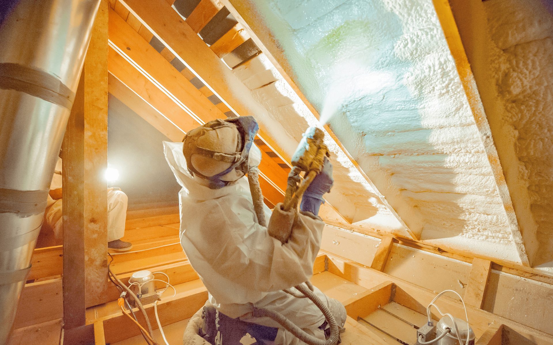 Worker in protective suit applying spray foam insulation to the wooden beams of an attic ceiling.