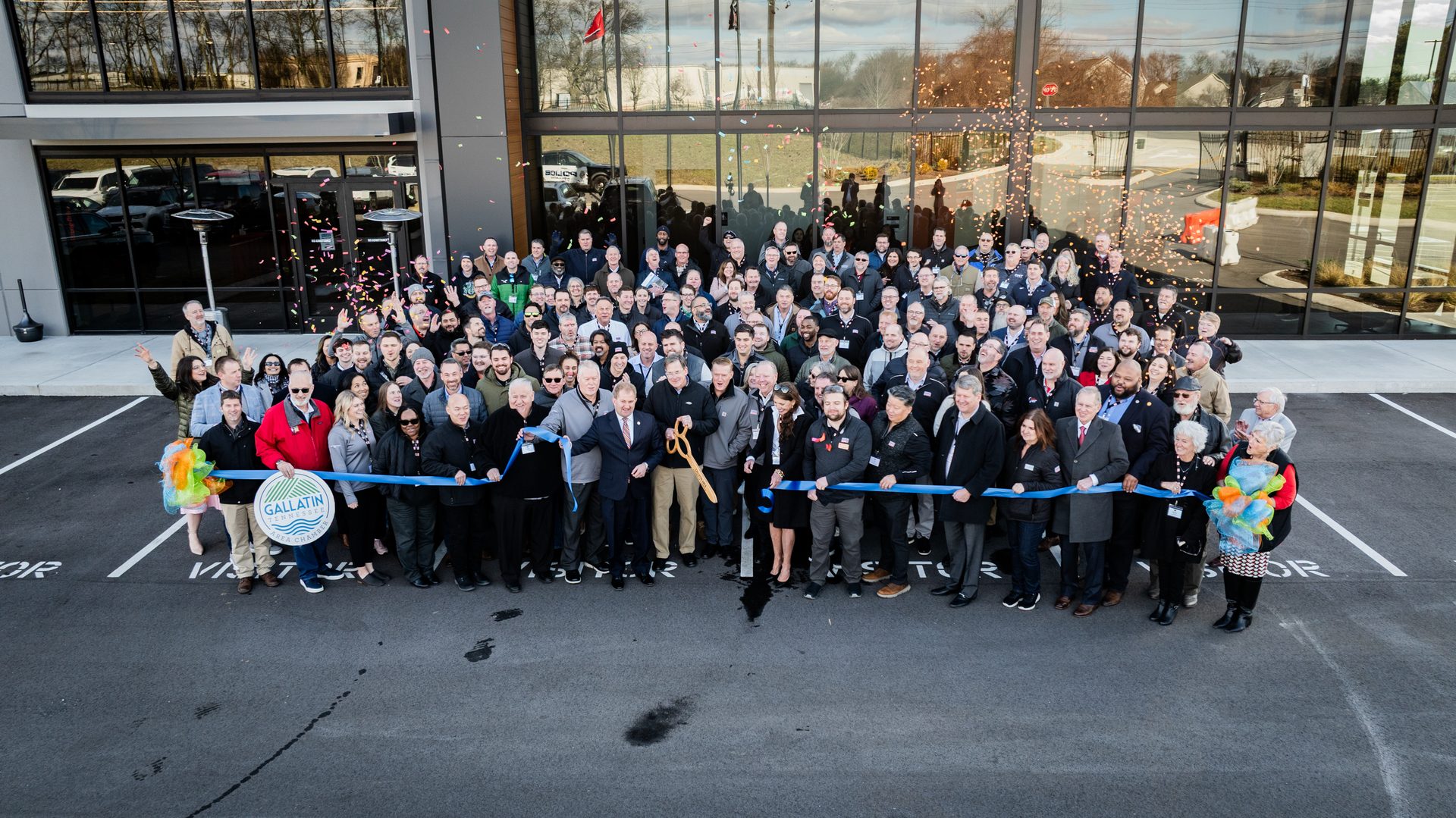 A large group celebrates a ribbon-cutting ceremony in front of a modern building with confetti.