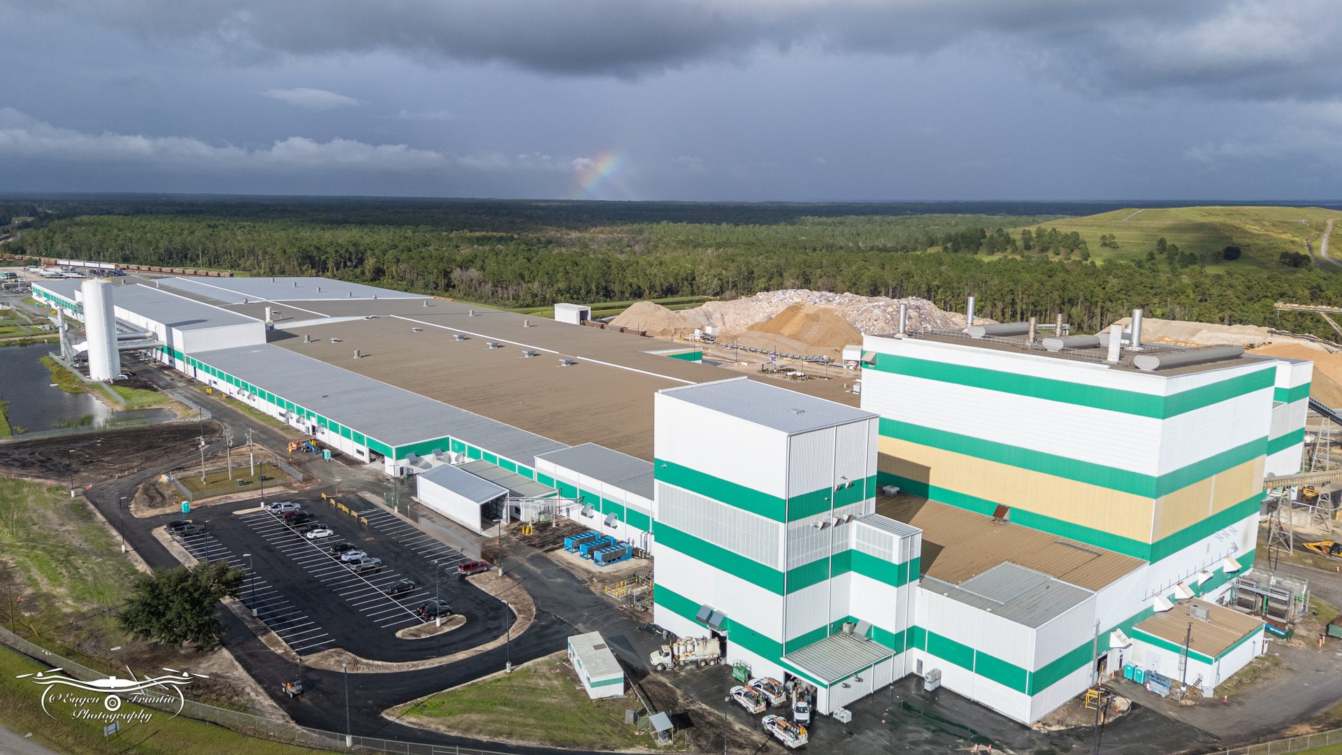 Aerial view of a large industrial facility with white and green buildings, vast forests, and a faint rainbow.