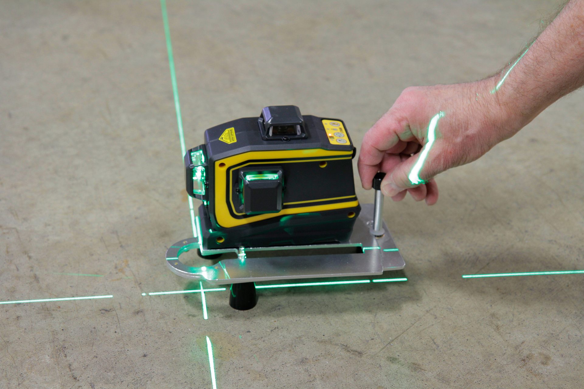 A hand adjusts a laser level emitting green lines onto the floor and the hand.