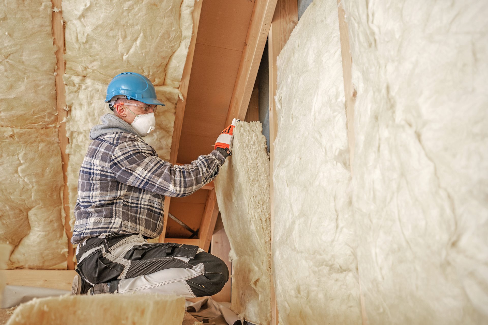 Worker installing insulation in an attic, wearing a hard hat, mask, and gloves.