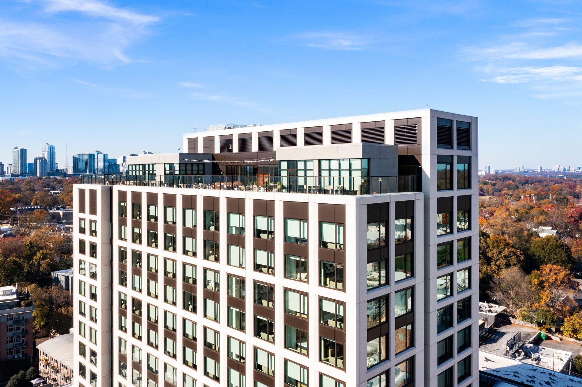 Modern high-rise building with a rooftop terrace, autumn trees, and a distant city skyline.
