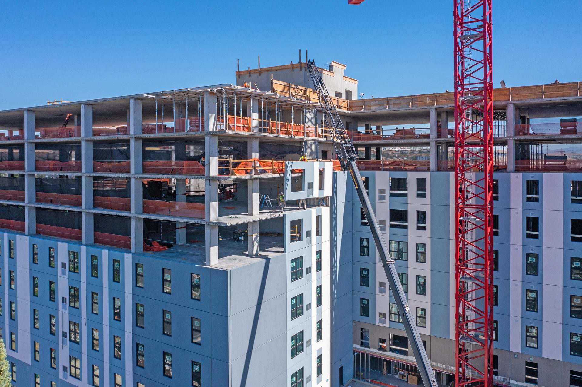 A multi-story building under construction, a crane lifting a wall panel with workers on site.
