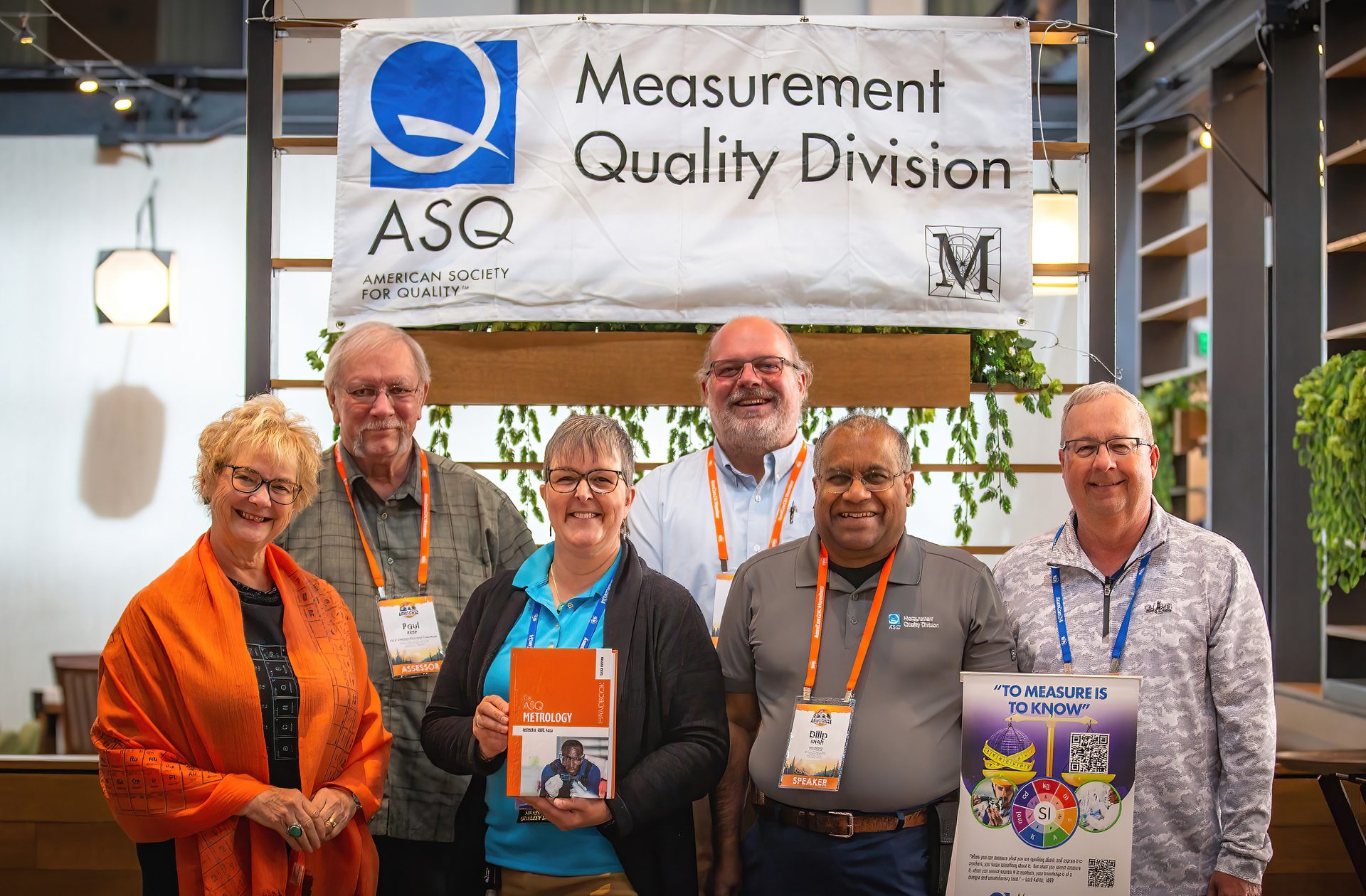 Six smiling members of the ASQ Measurement Quality Division posing for a group photo under their banner.
