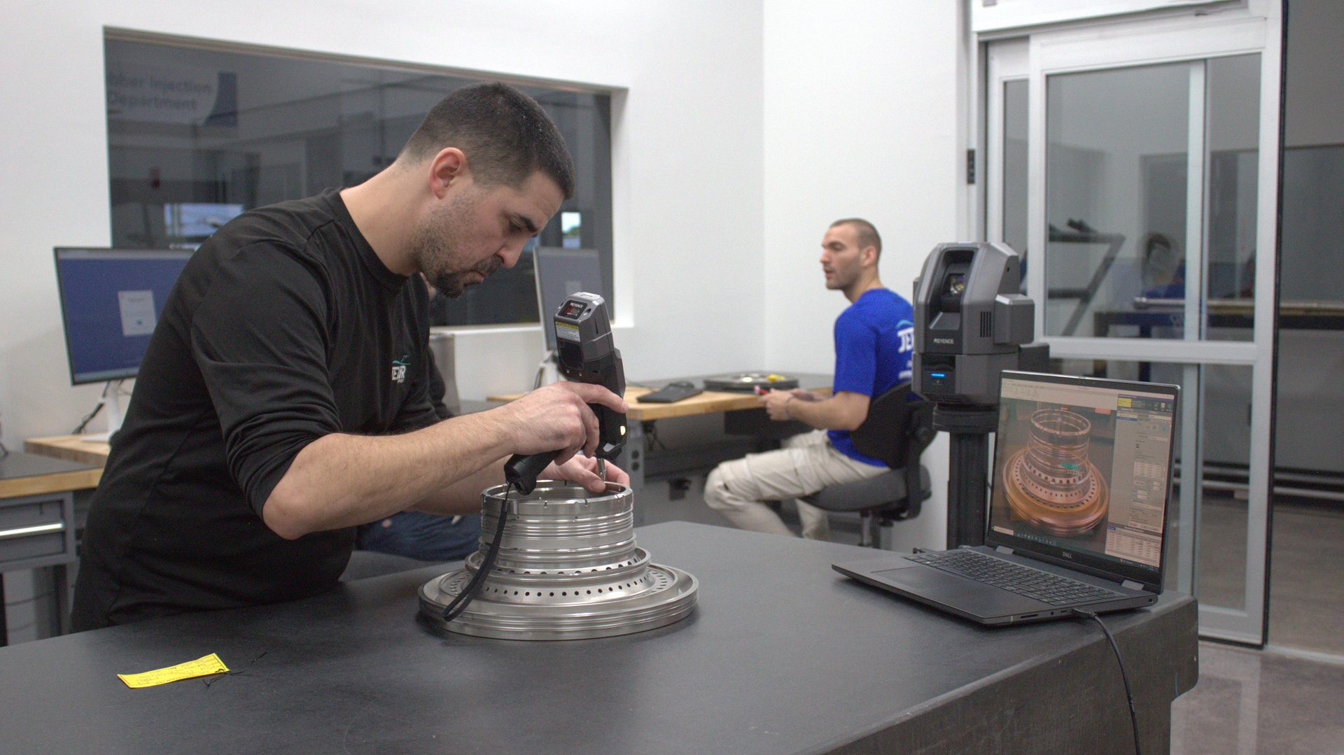 Man scanning a complex metal part with a handheld device; laptop shows its 3D model.
