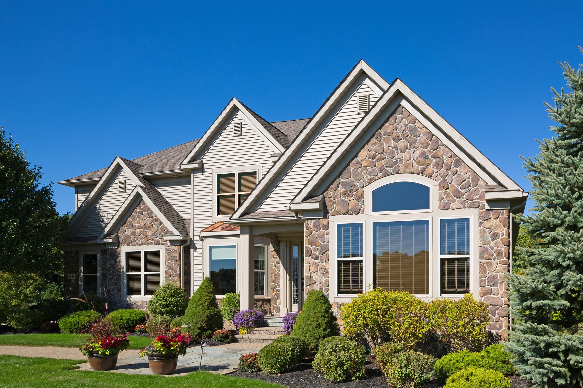 A picturesque two-story house featuring stone and siding, surrounded by vibrant plants and a clear blue sky.