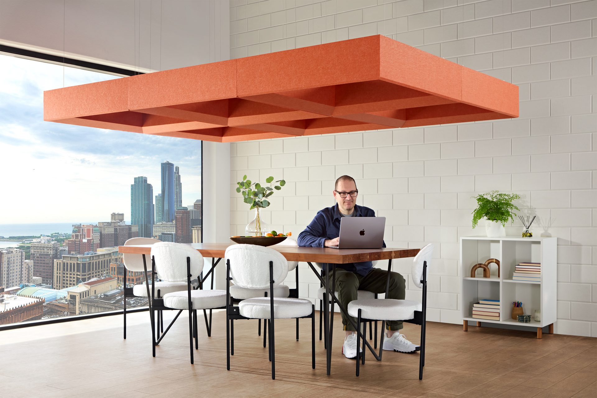 Modern workspace with a man on a laptop, an orange acoustic panel, city view, and white chairs.