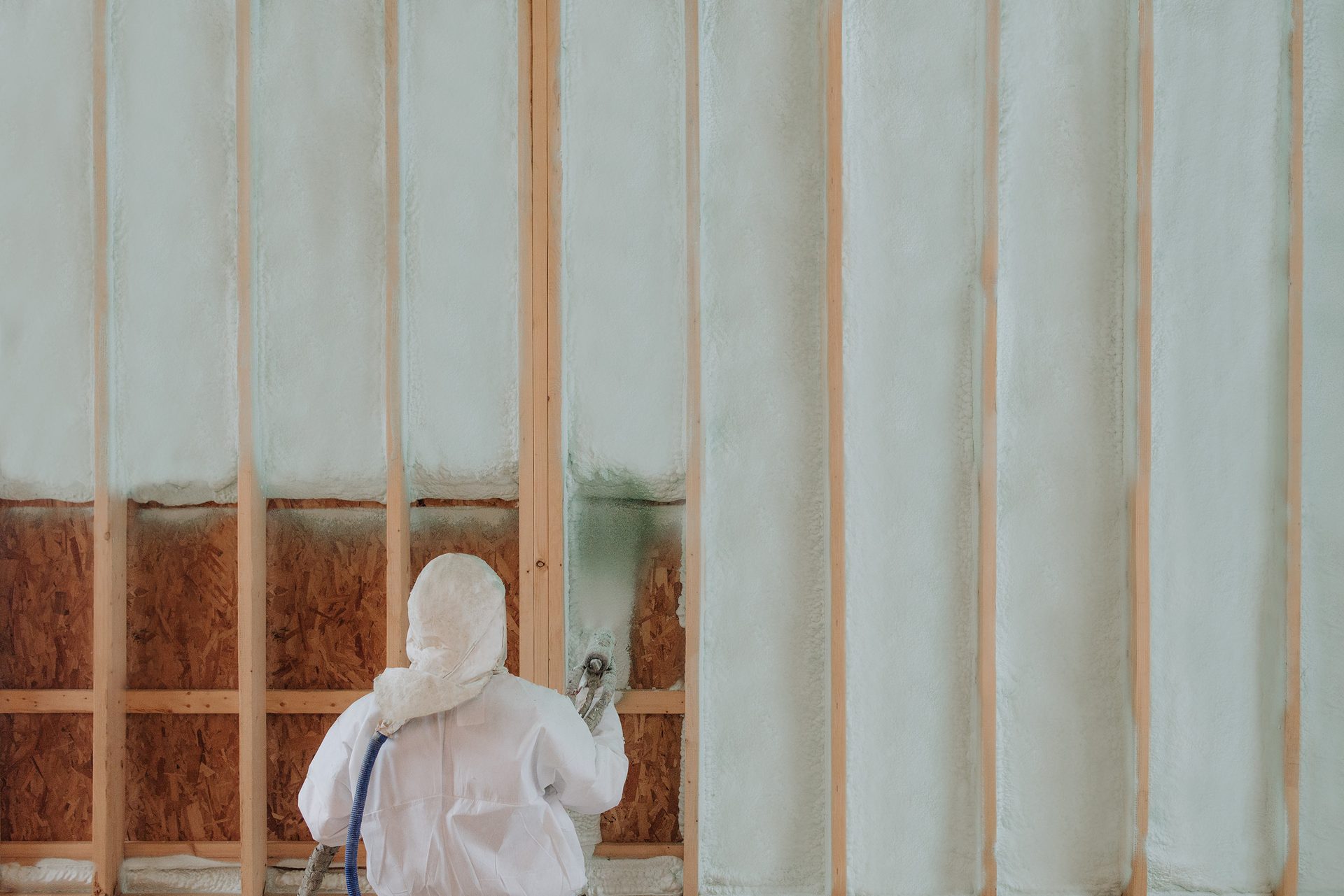 Worker in protective suit spraying foam insulation into wall framing.