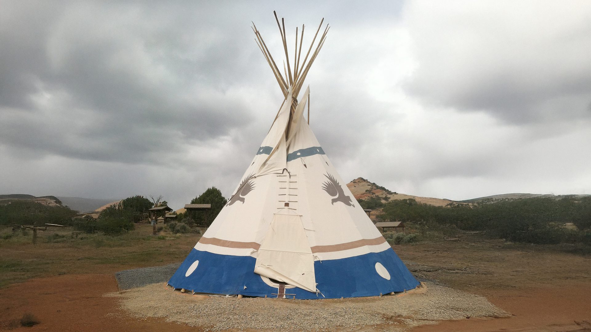 Decorated tipi with blue base, eagle art, in landscape under cloudy sky.