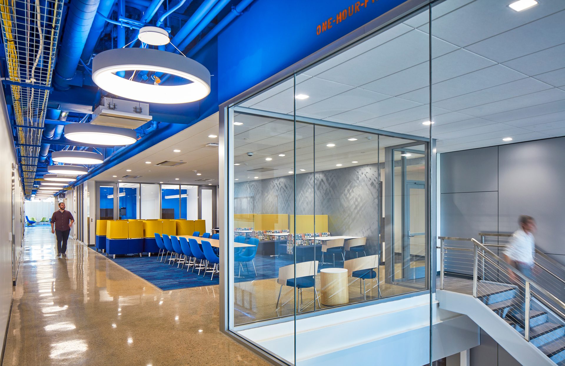 Bright office with blue ceilings, yellow furniture, glass walls, and a person walking.