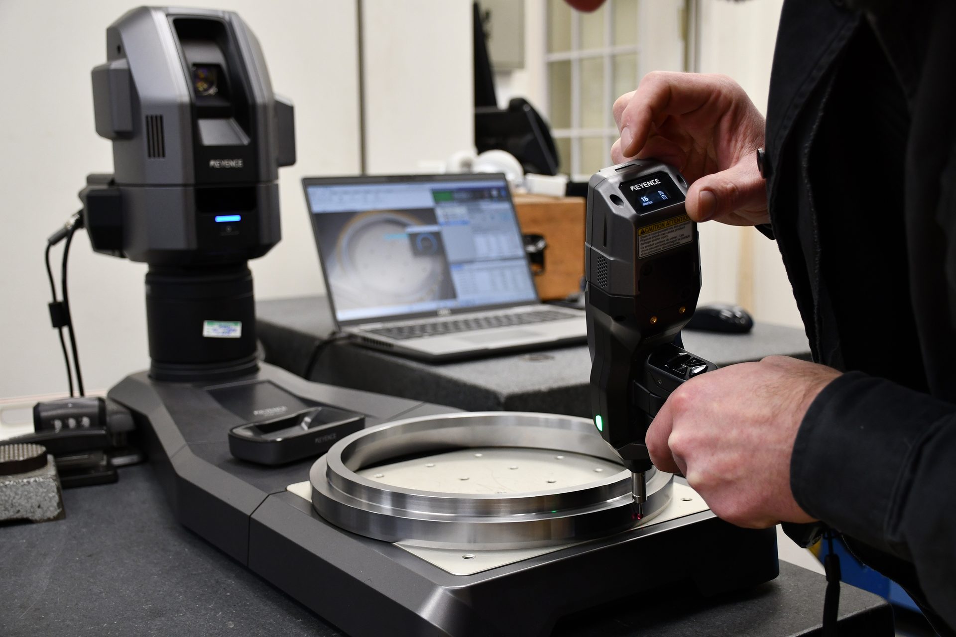 A person uses a handheld Keyence device to measure a metal ring on a precision measurement system.
