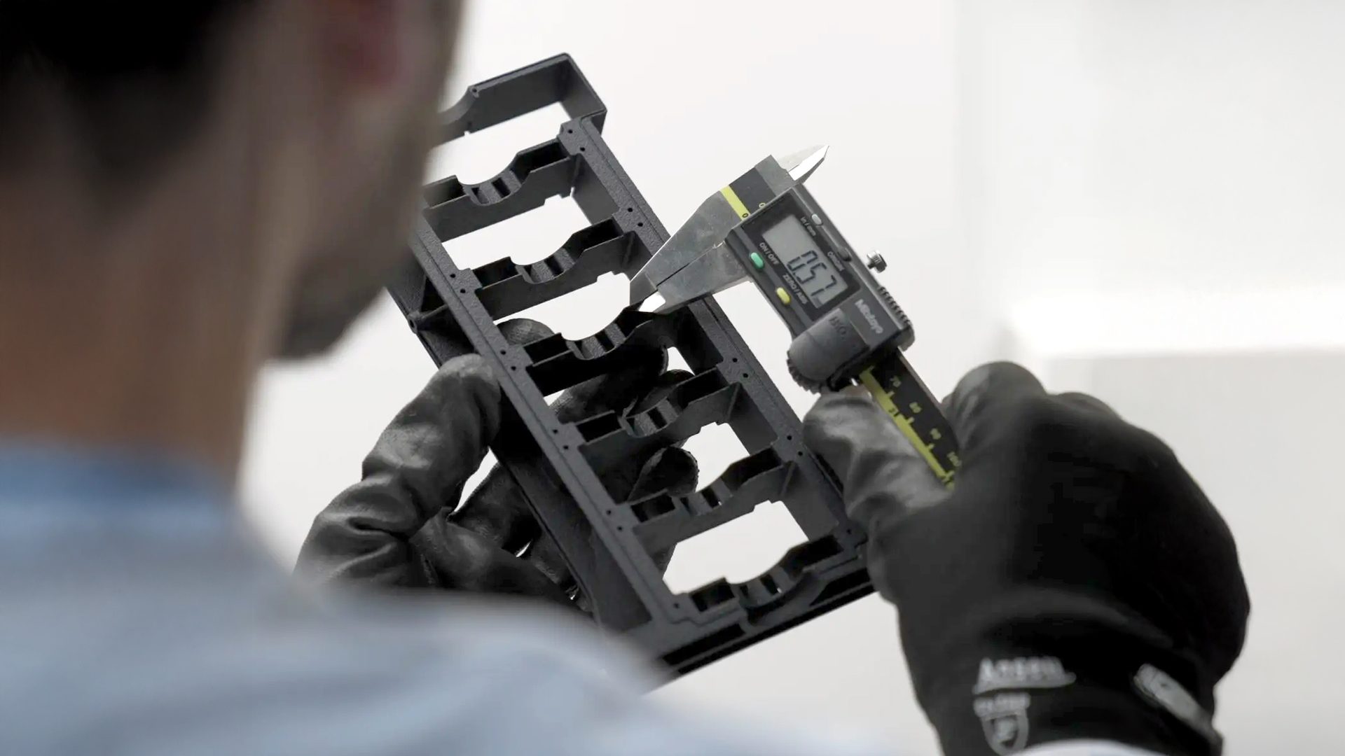 A person in black gloves measures a black component with a digital caliper.