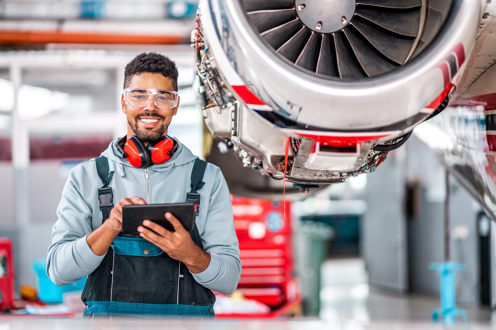 Smiling mechanic in safety gear with a tablet next to a jet engine in a hangar.