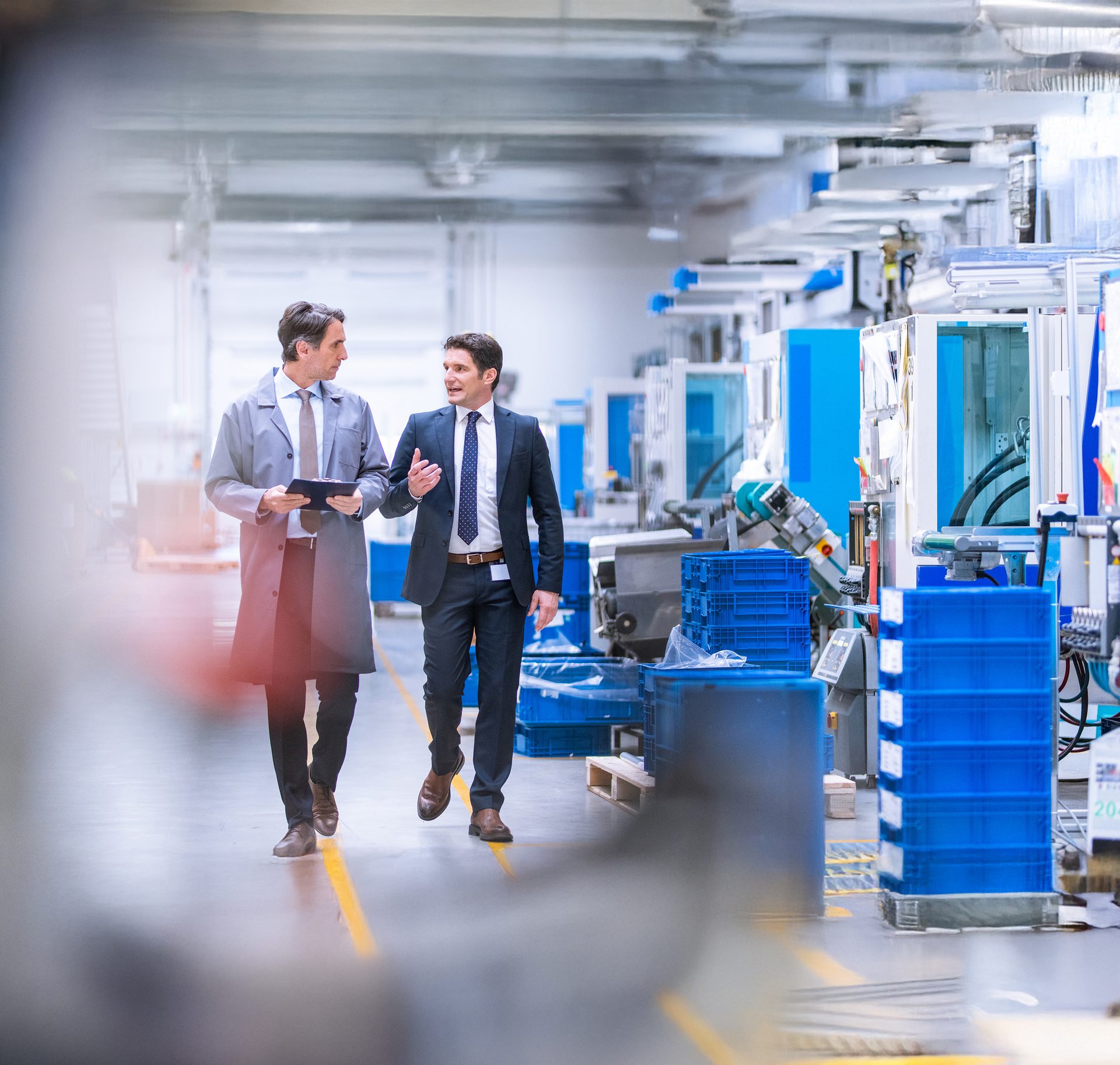 Two men, one in a lab coat and one in a suit, walk and talk in a factory.