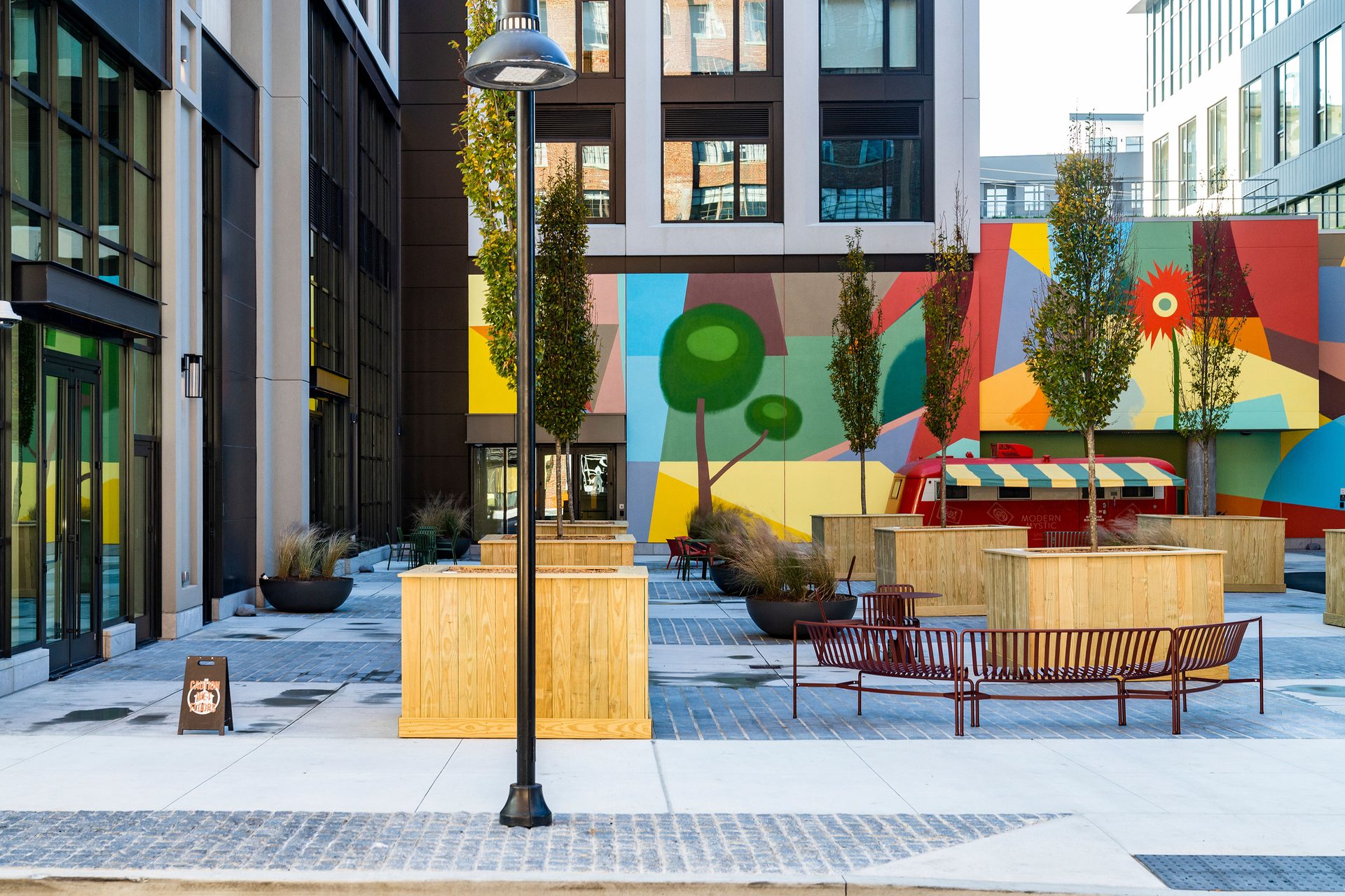 Vibrant urban courtyard with a colorful mural, modern buildings, trees, a red food truck, and benches.