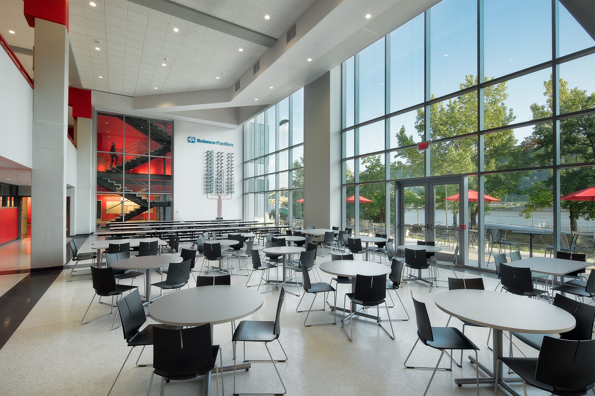 Bright, modern dining hall with river views, floor-to-ceiling windows, tables, and a red staircase.