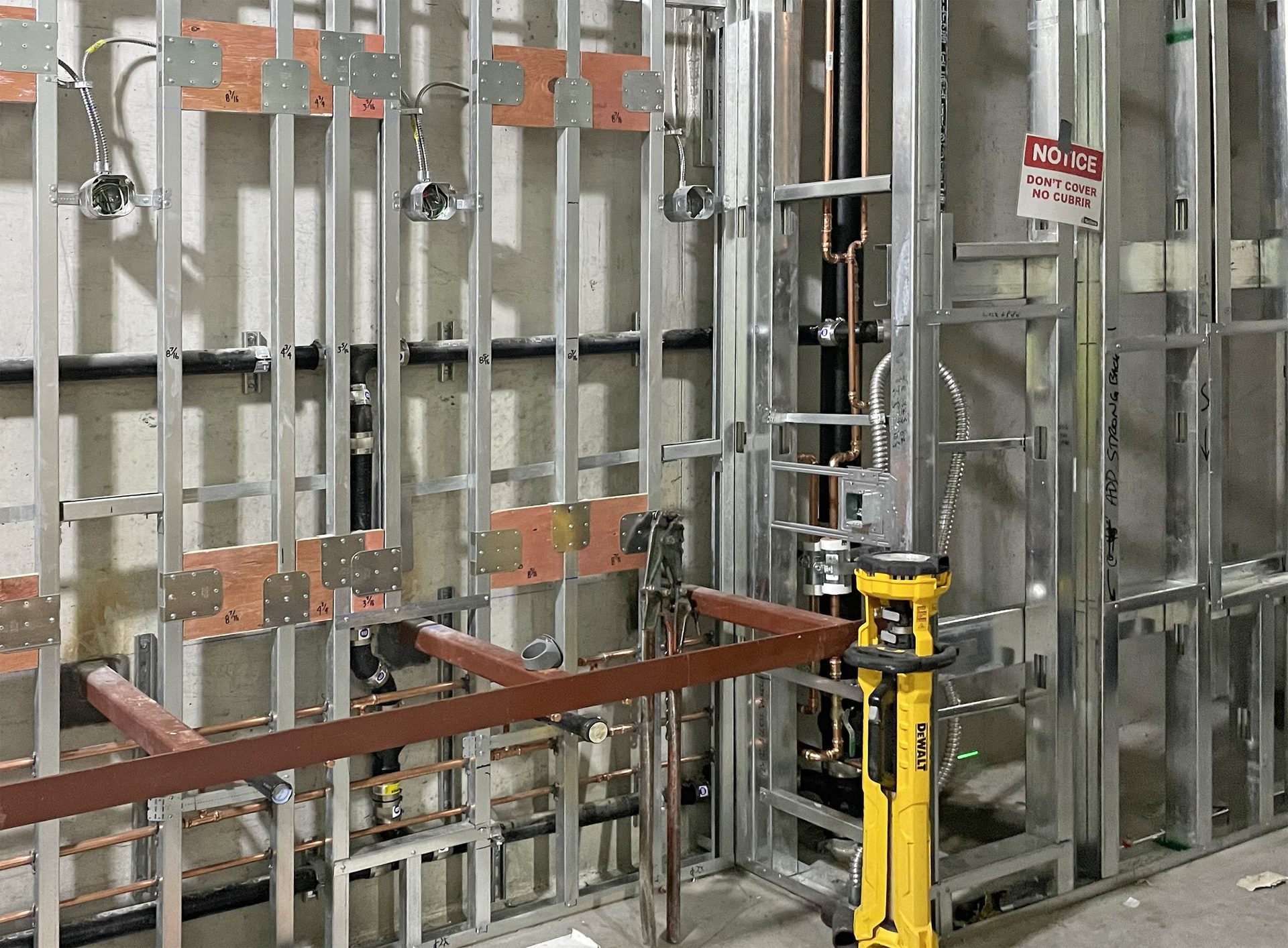 Metal stud wall under construction with electrical wiring, plumbing, wood blocking, and a "DON'T COVER" sign.