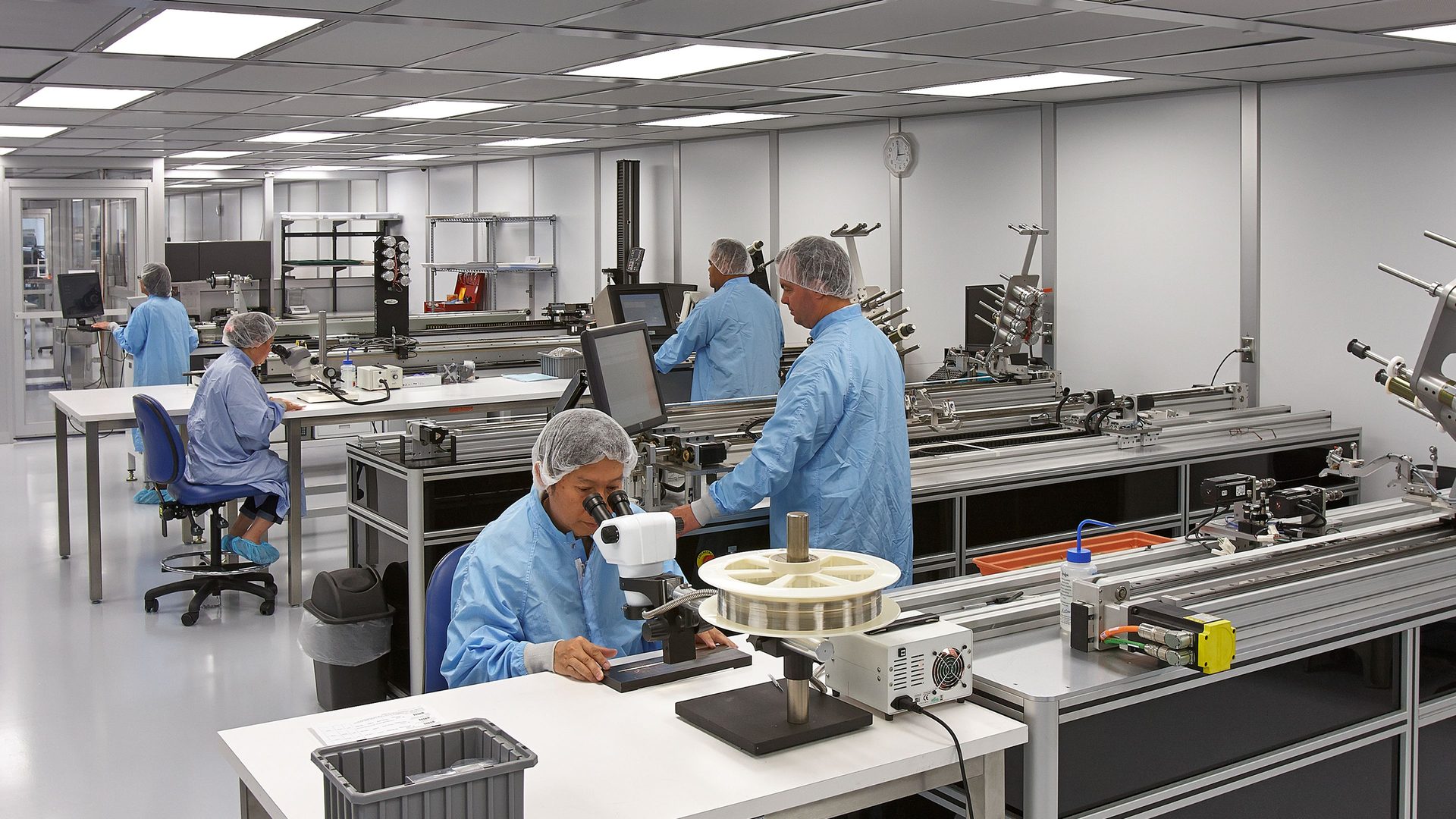 Workers in blue cleanroom suits and hairnets operate machines and microscopes in a clean lab.