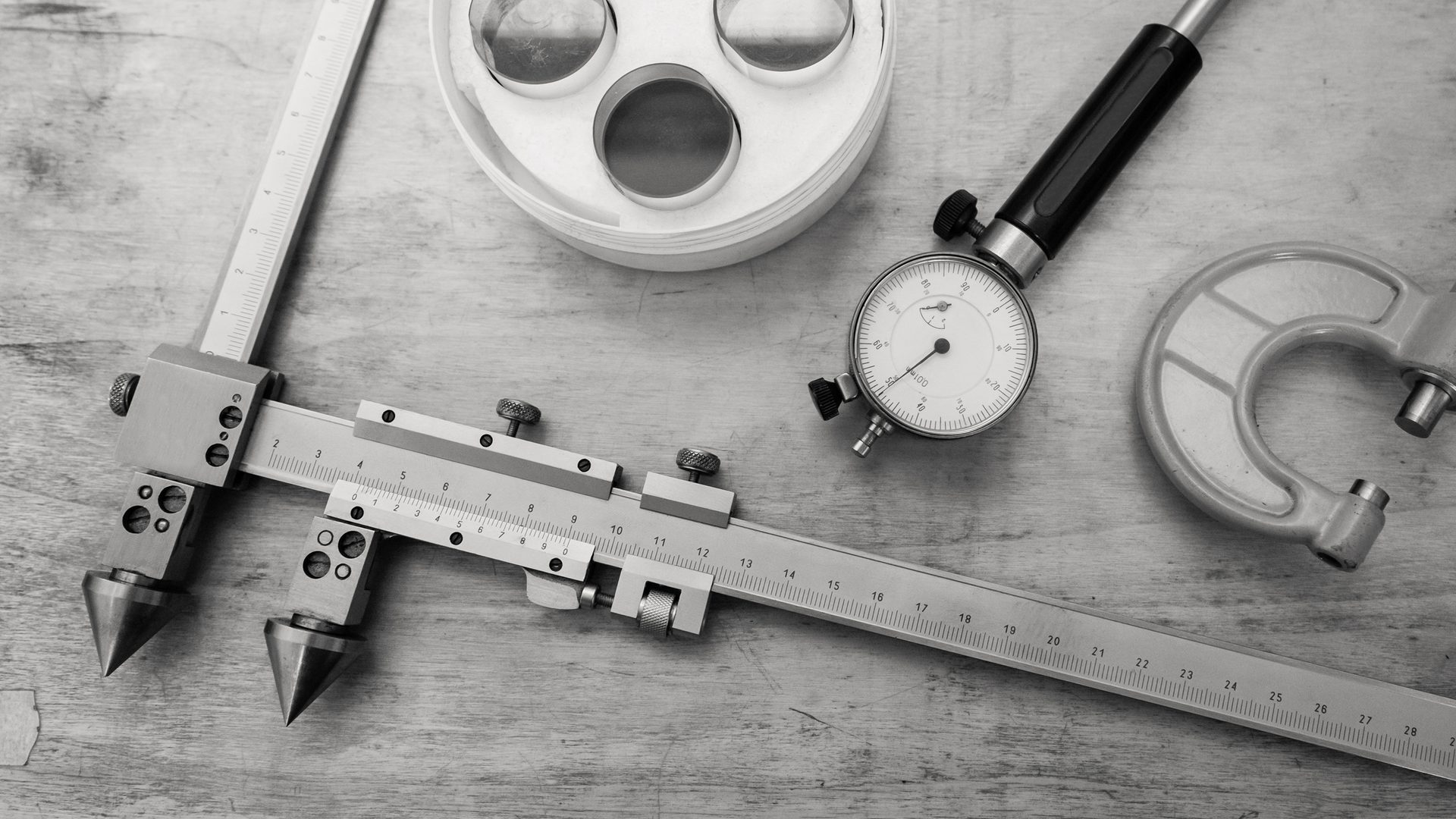 Precision measuring tools: calipers, micrometer, dial indicator, and lenses on wood.