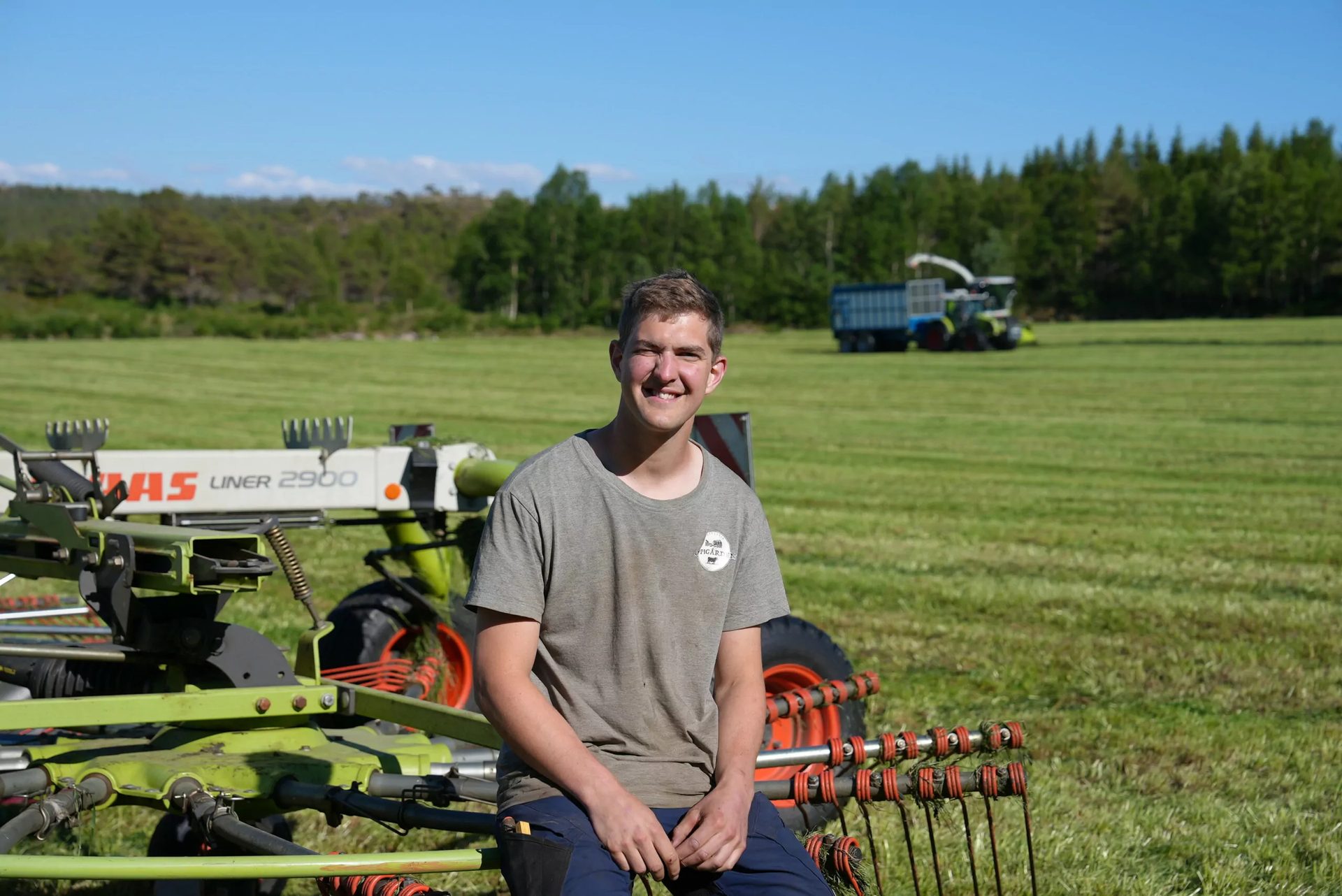 Smiling young man on farm equipment in a green field with a tractor and trees.