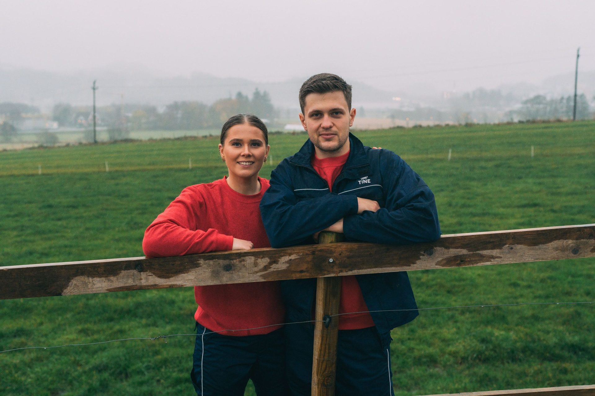 A woman and man lean on a wooden fence in a misty, green field.